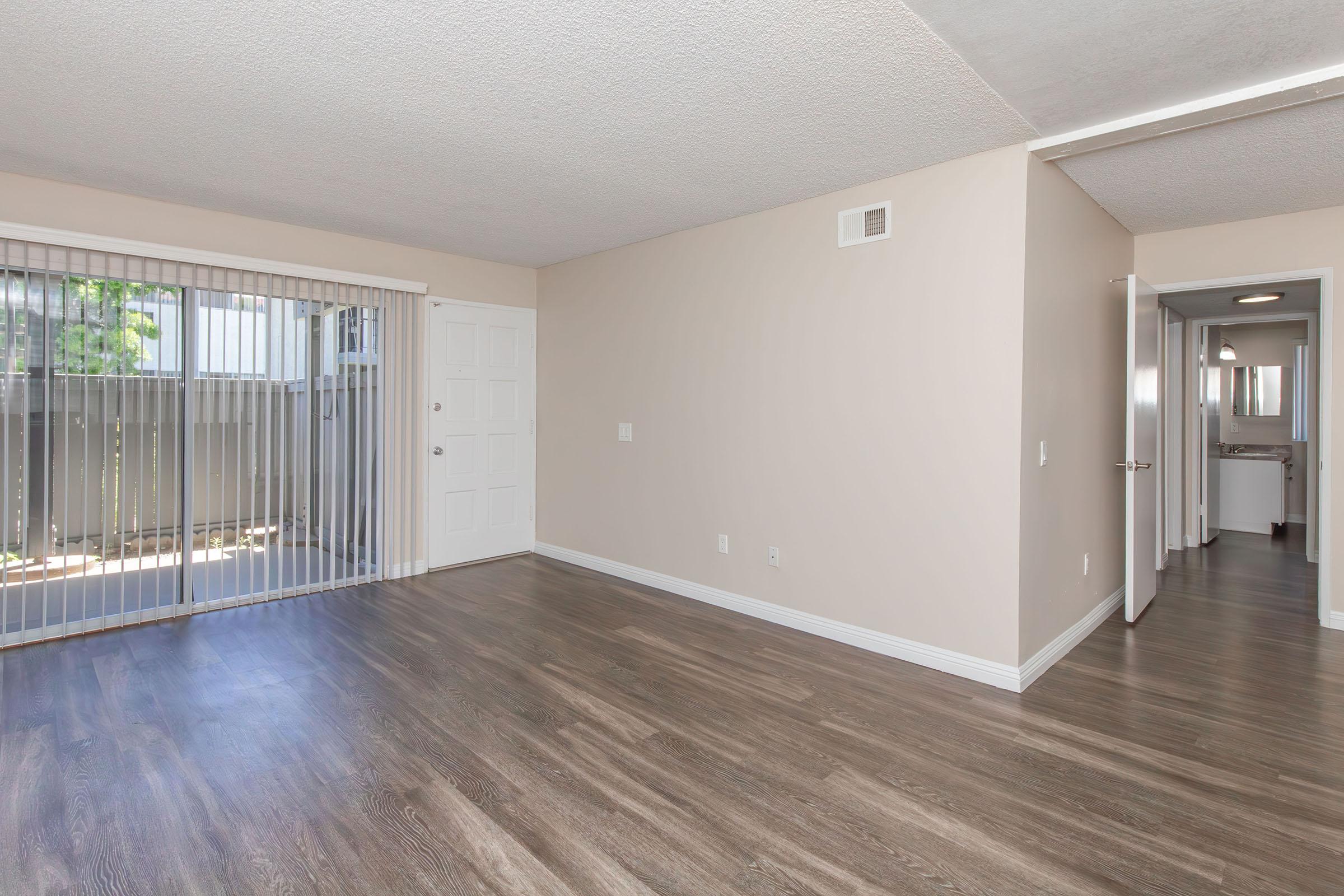 A spacious and well-lit room with light-colored walls and laminate flooring. It features a sliding glass door leading to an outdoor space, and an adjacent doorway that leads to a bathroom. The overall ambiance is clean and modern, ideal for living or entertaining.