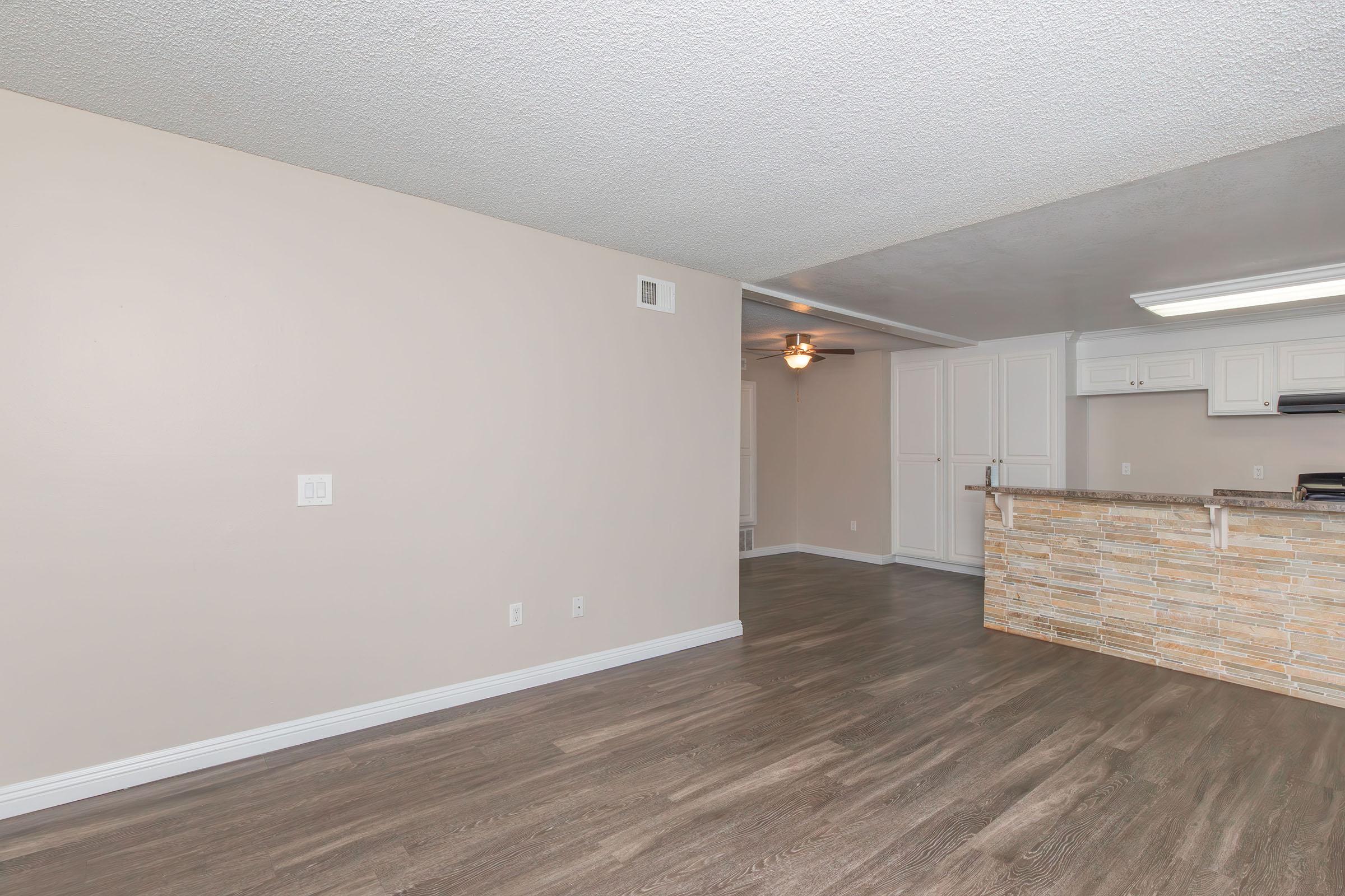 Spacious living area with light-colored walls, a textured ceiling, and wood-style flooring. To the right, a modern kitchen with a breakfast bar featuring stone accents. A ceiling fan is visible in the background, enhancing the open layout. Ideal for cozy living and entertaining.