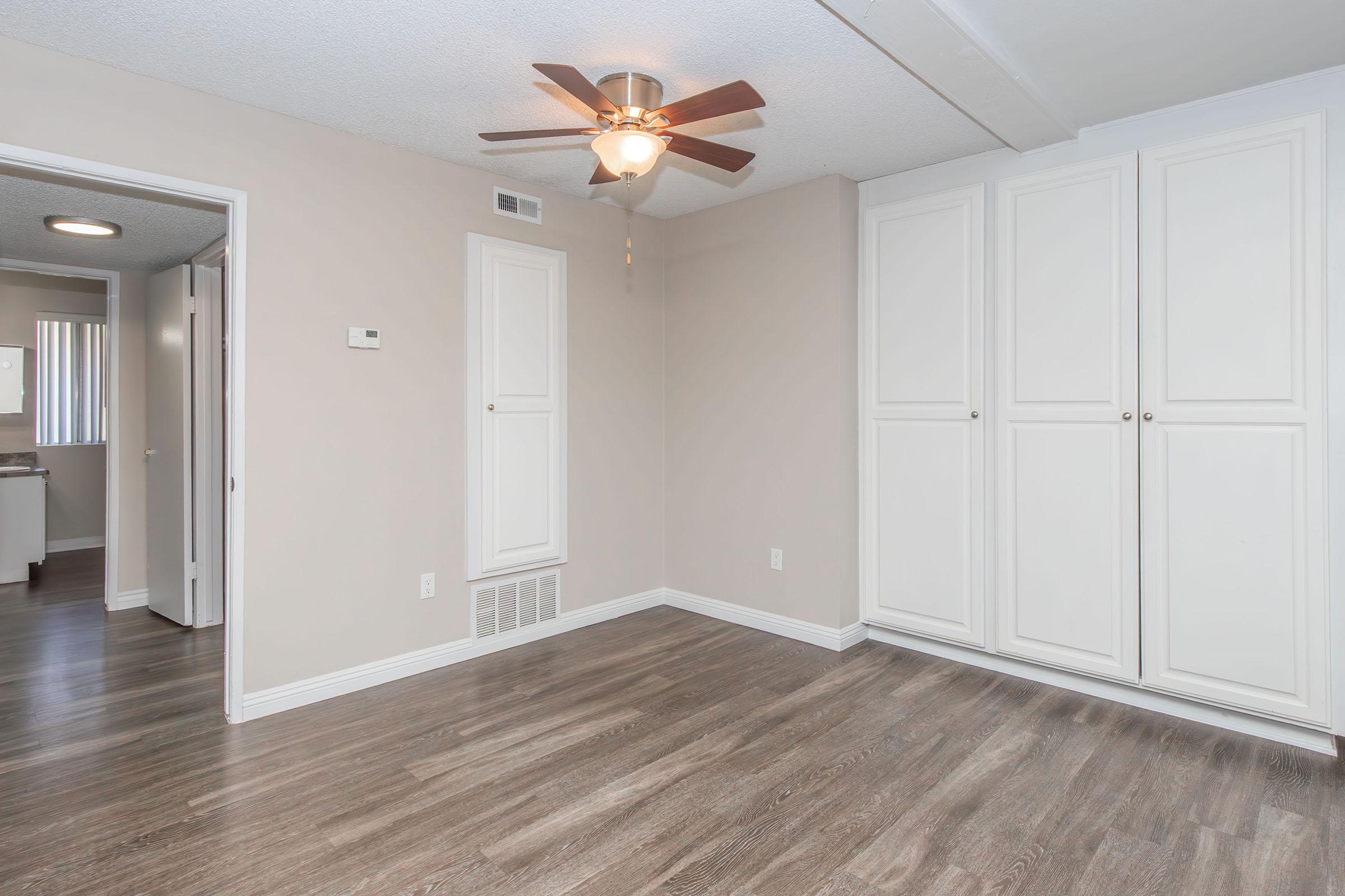 A well-lit room featuring a ceiling fan, light-colored walls, and modern laminate flooring. One wall has built-in cabinets with white doors, and there's a doorway leading to another room. A thermostat is mounted on the wall, and a light fixture is visible in the ceiling.