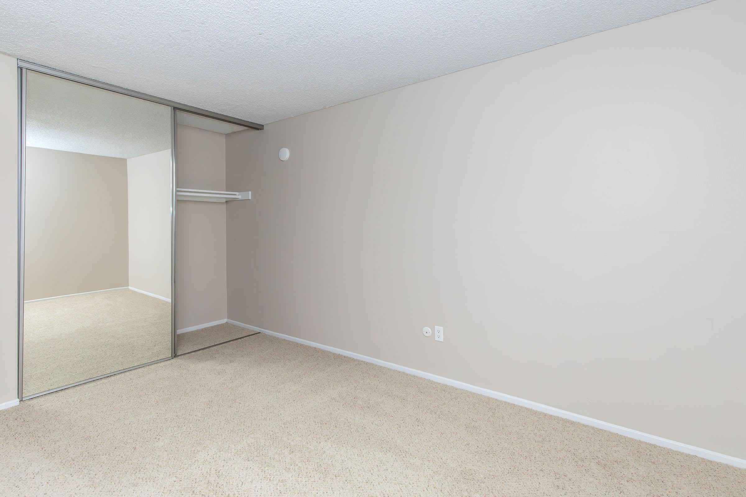 Empty room with light beige walls and carpeting. A floor-to-ceiling mirror is located on one wall, reflecting part of the room. There is a shelf and a light fixture on the ceiling. The space appears clean and minimalistic, suitable for various uses or decoration.