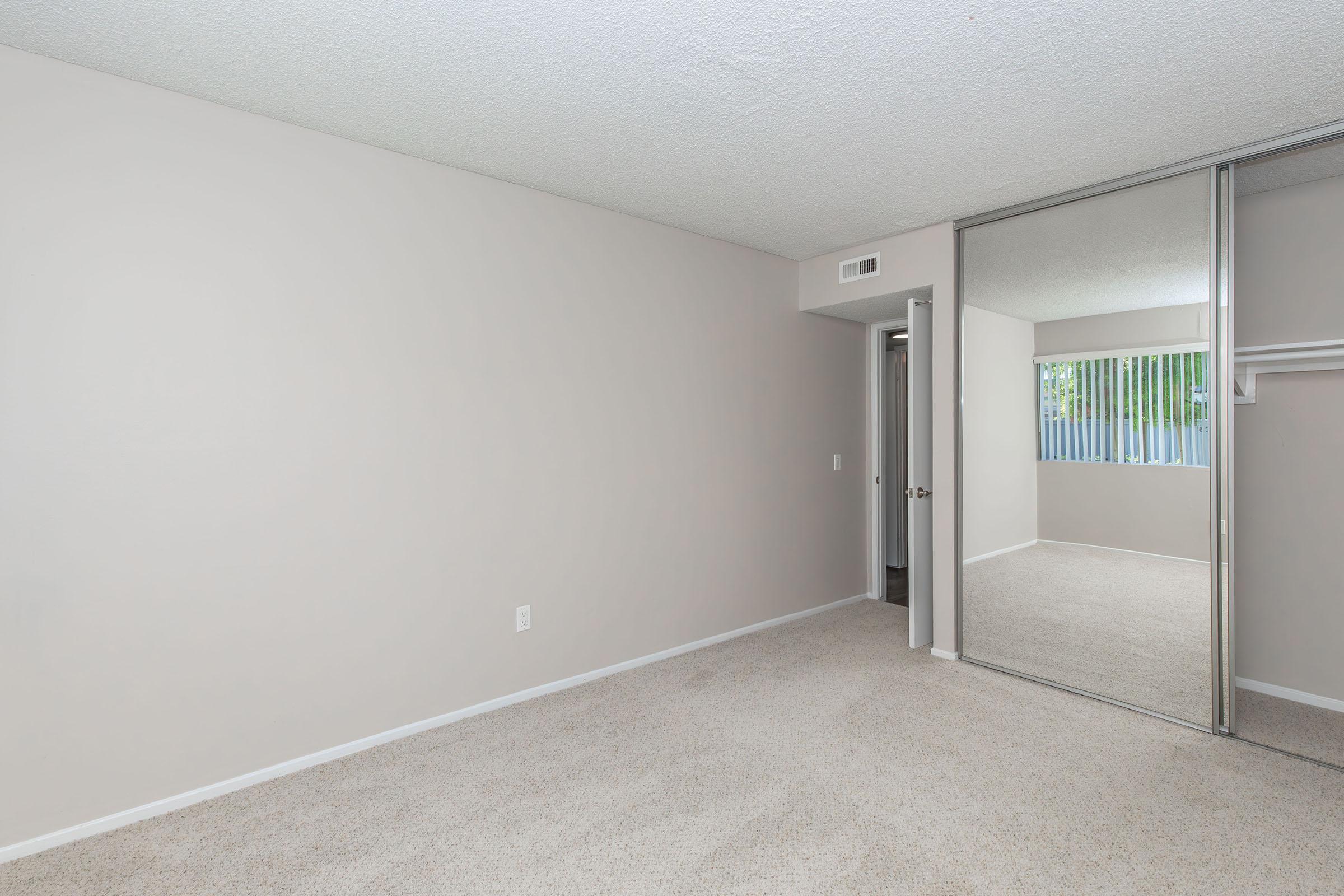 Empty room with light gray walls, carpeted floor, and a large mirrored closet on one side. A doorway leads to another space, and there's a window with blinds, providing natural light. The overall atmosphere is clean and spacious, with a neutral color palette.
