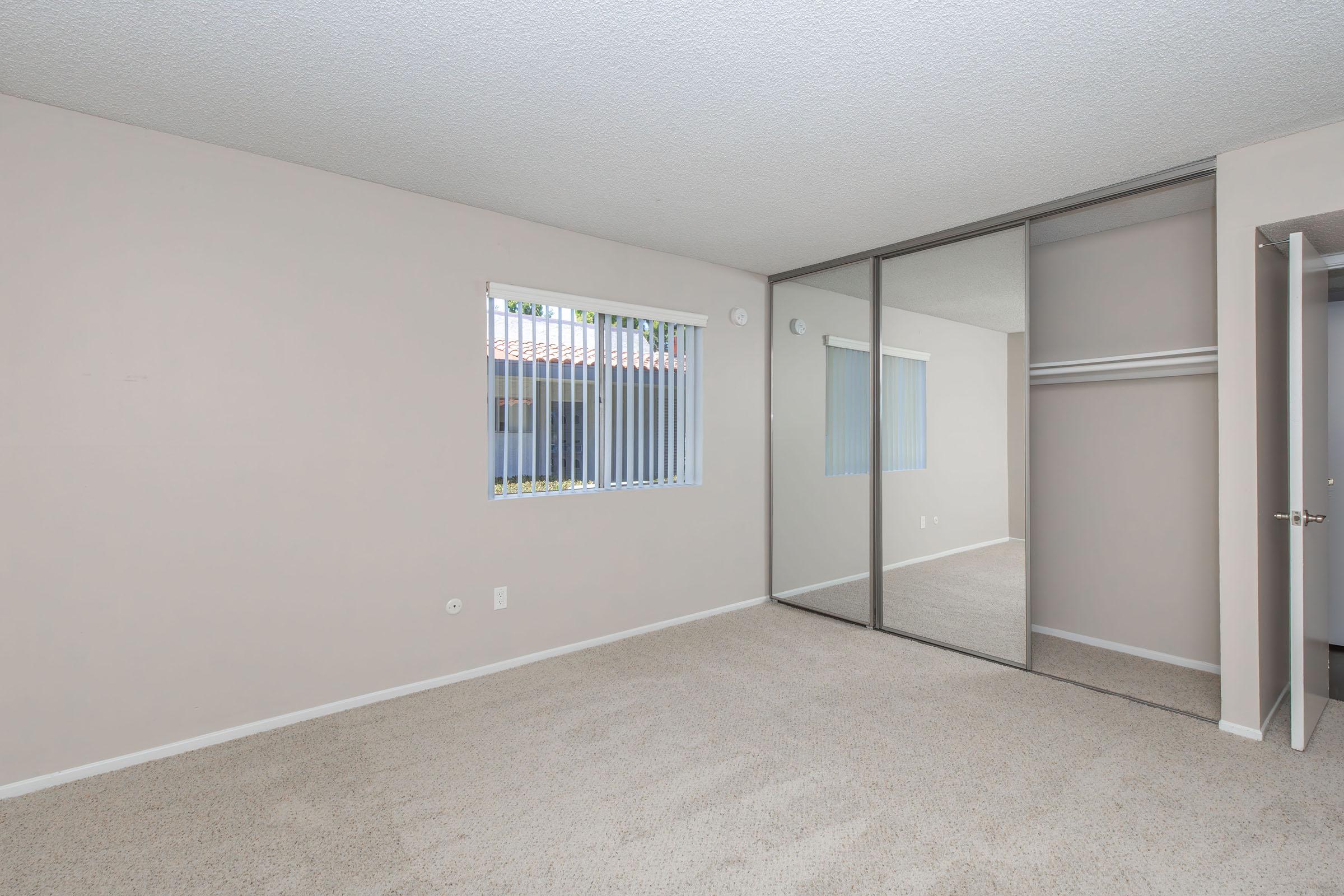 Empty room with beige walls and carpet, featuring a window with vertical blinds. There is a mirrored closet on one side, and a door partially open leading to another space. The overall ambiance is bright and neutral.