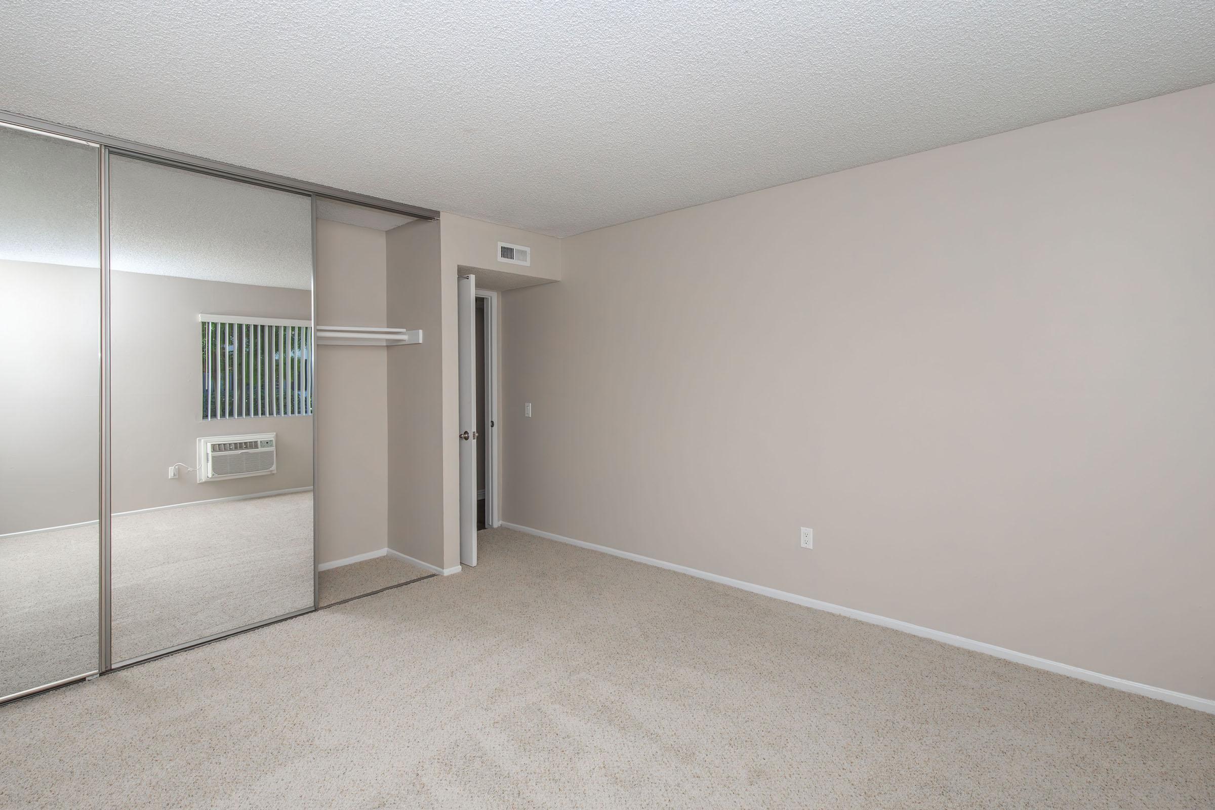 Empty room with beige walls and carpet. Features a mirrored closet on the left, a small air conditioning unit under a window, and a doorway leading to another space. Light from the window illuminates the room, creating a bright and neutral atmosphere.