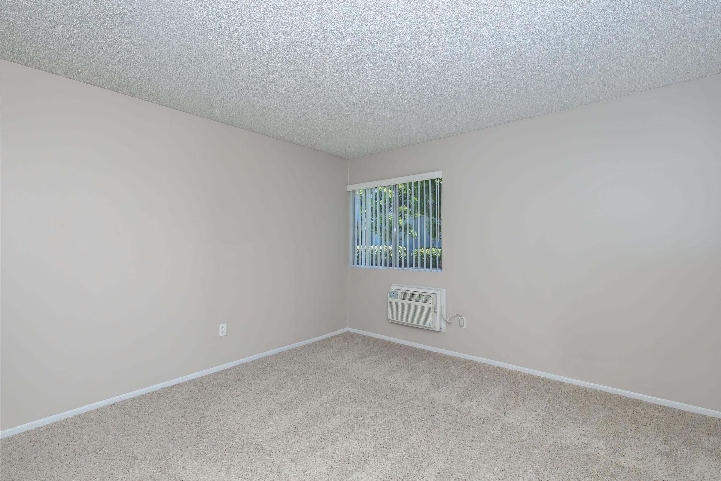 A vacant room with light beige walls and carpet. It features a small window with vertical blinds and an air conditioning unit installed beneath the window. The room is well-lit and has no furniture, giving it an open and spacious appearance.