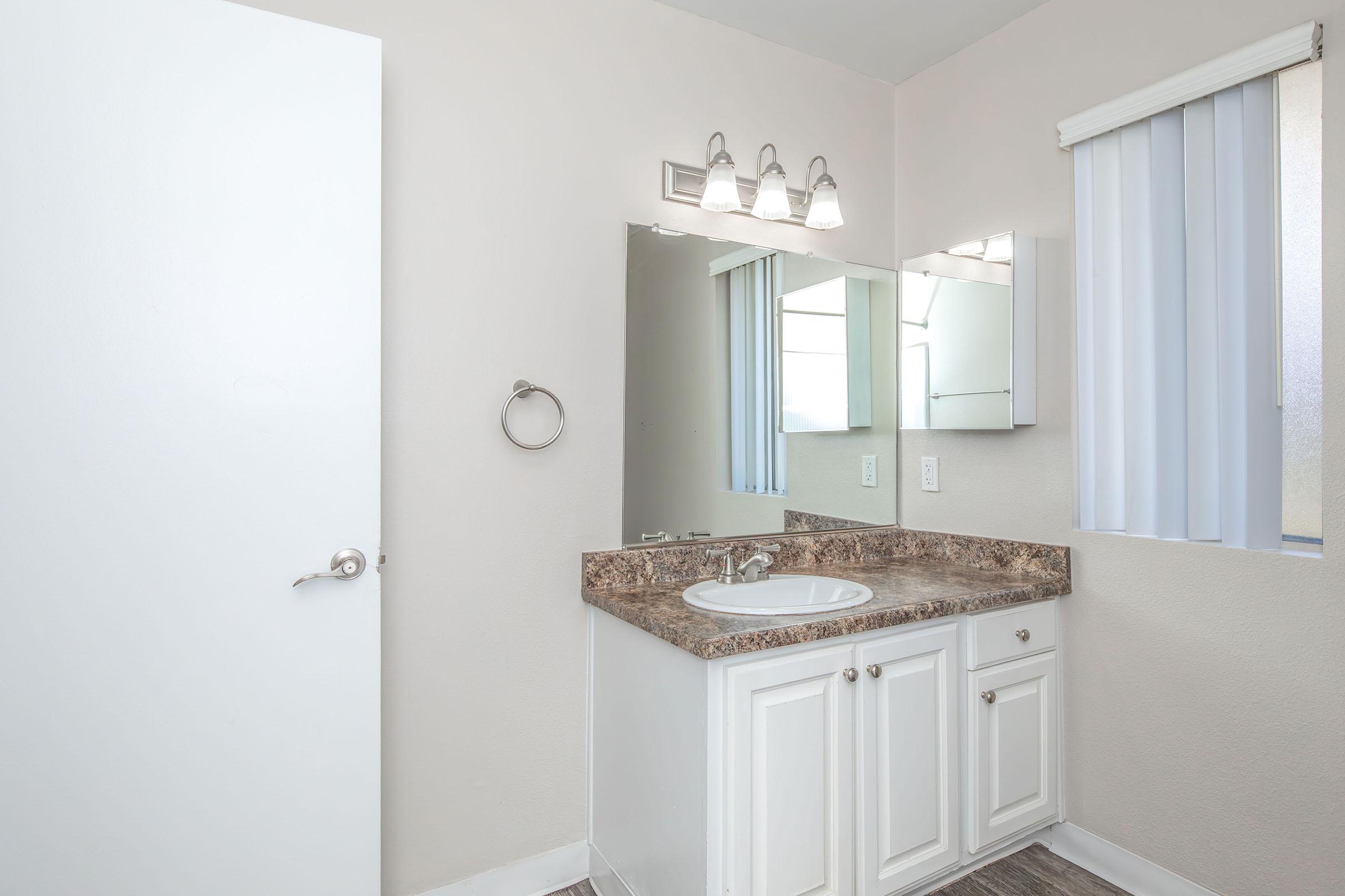 A modern bathroom featuring a white door, a countertop with a sink, and a large mirror. There are three light fixtures above the mirror, and the window is covered with vertical blinds, allowing natural light to enter the space. The walls are painted in a light shade, creating a clean and bright atmosphere.