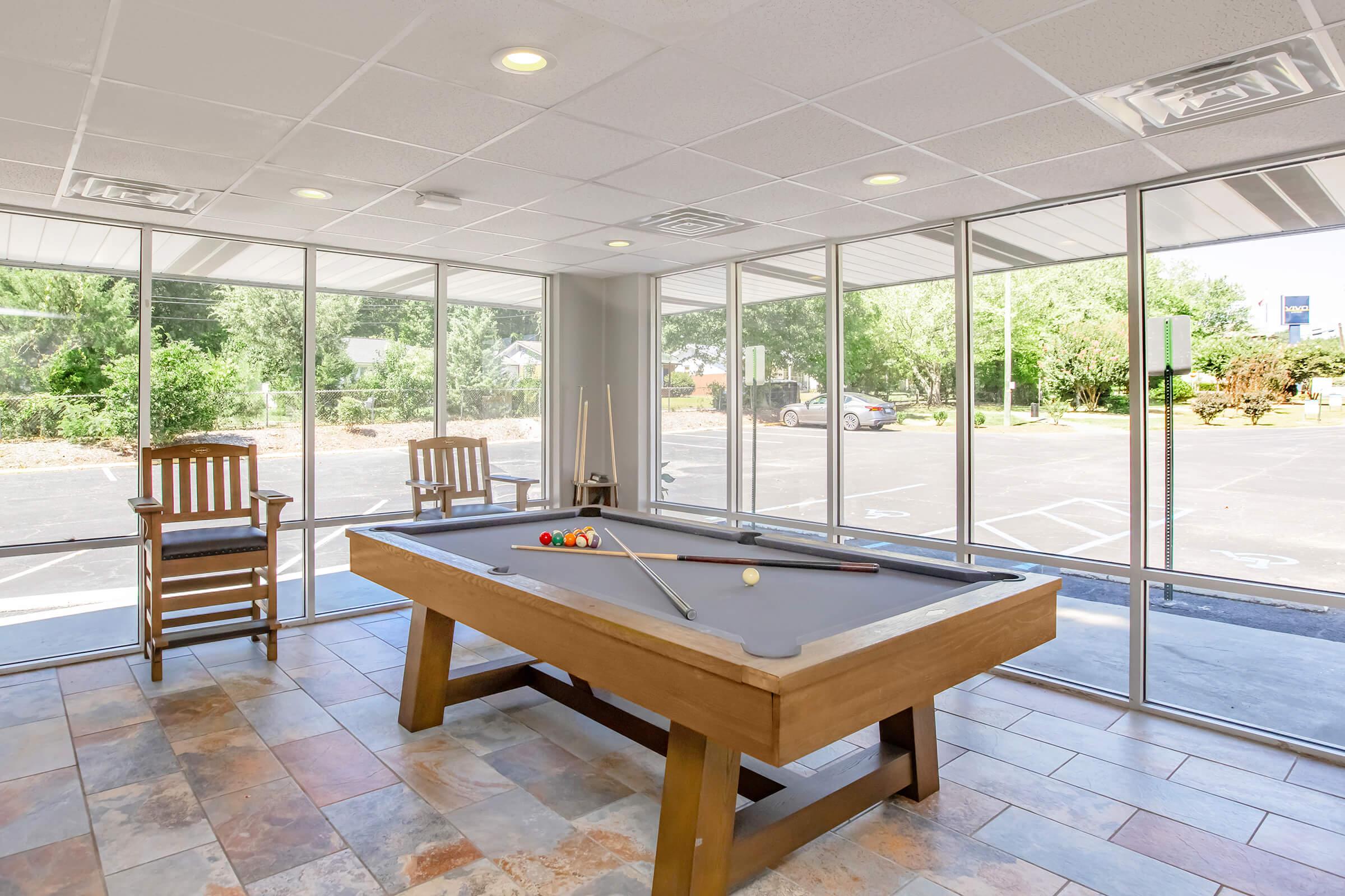 Bright and spacious game room featuring a billiards table with a cue stick and colorful pool balls. Large windows let in natural light, offering views of the outdoor area. Two wooden rocking chairs are positioned nearby, creating a welcoming atmosphere for relaxation and leisure.