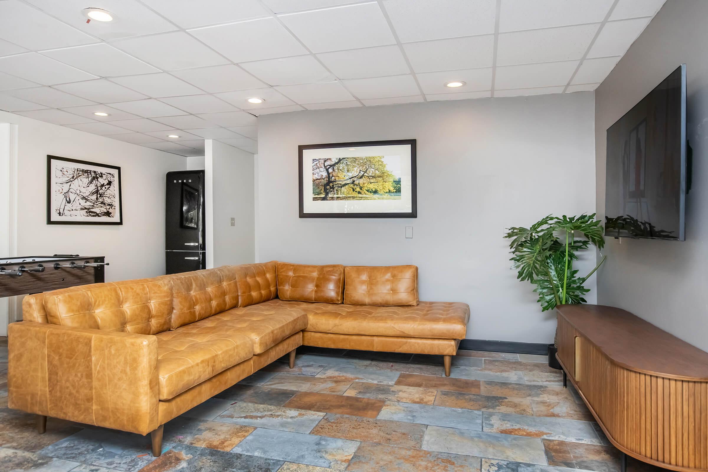 A modern living room featuring a brown leather sectional sofa, a wooden TV cabinet, and a large flat-screen TV mounted on the wall. There is a potted plant in the corner and artwork displayed above the sofa. The floor is made of stone tiles, and the walls are painted in neutral colors.