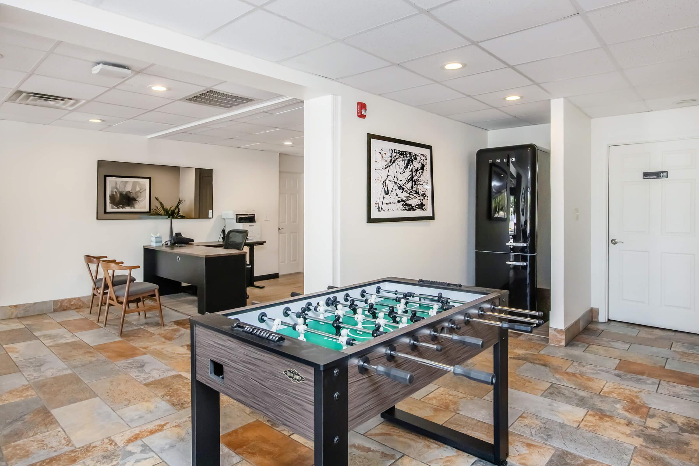 A spacious game room featuring a foosball table in the foreground. In the background, there is a modern kitchen area with a refrigerator, and four wooden chairs at a table. The floor is tiled, and the walls feature artwork. The room is brightly lit with overhead lighting.