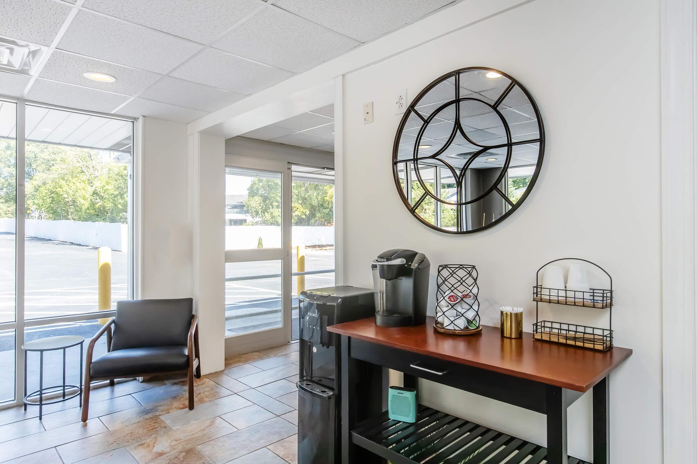 A bright reception area with large windows letting in natural light. There's a dark wood console table with a coffee maker, a wire basket with supplies, and decorative items. A round mirror hangs on the wall, and a modern black chair sits in the corner near the entrance, creating a welcoming atmosphere.