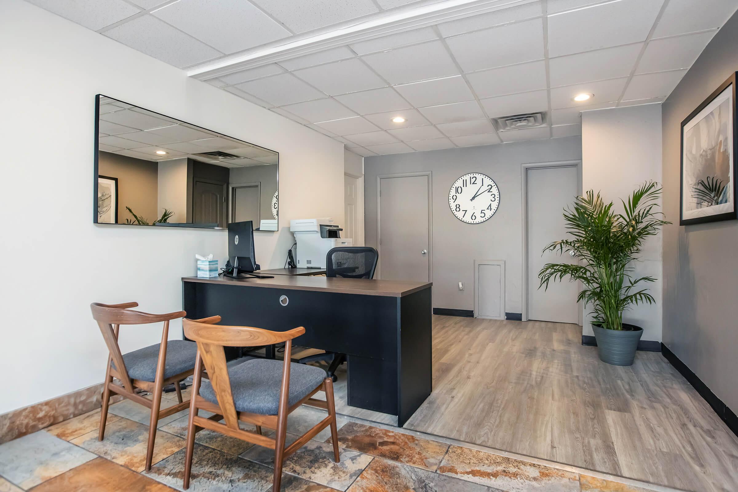 A modern office reception area featuring a black desk with two wooden chairs, a large mirror on the wall, and a potted plant. There is a clock displaying the time on the wall, and the floor is a mix of tile and laminate. The space is well-lit and has a minimalist design.