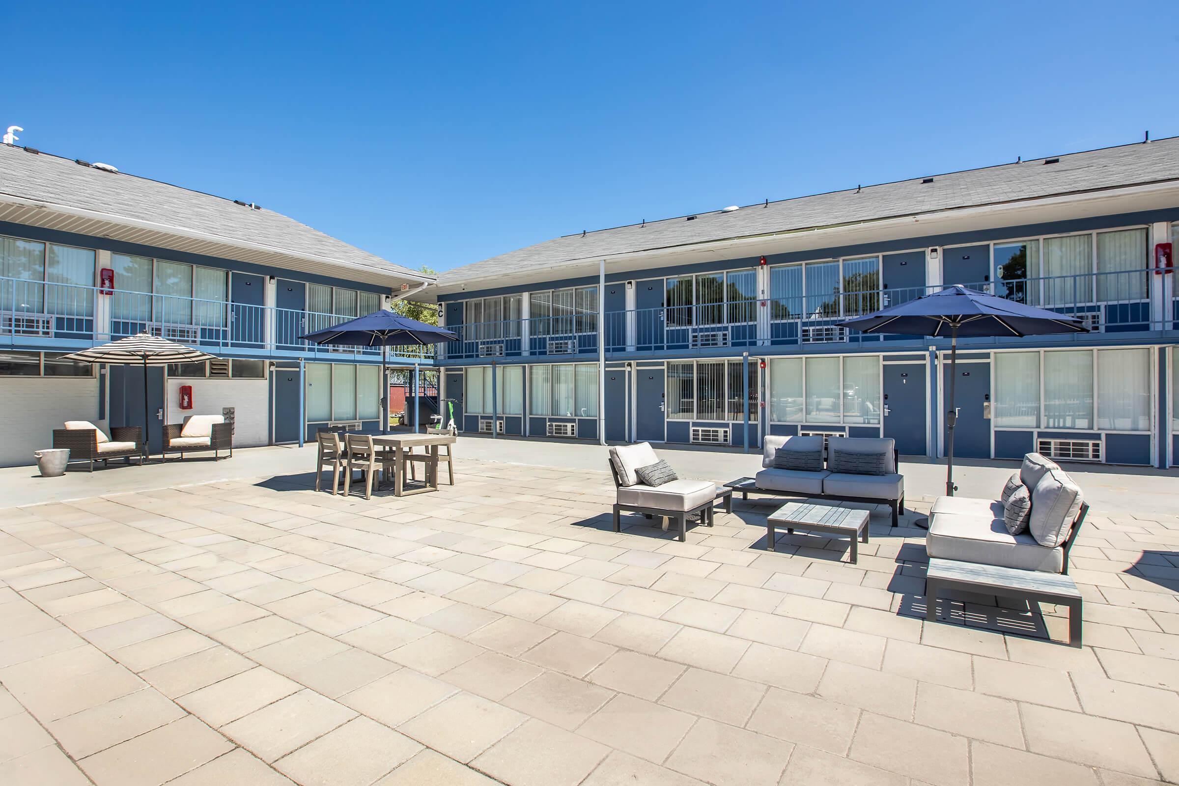 A courtyard of a motel featuring several outdoor seating areas with comfortable lounge chairs, a dining table, and umbrellas. The buildings are blue with white railings, and the setting is bright and sunny, ideal for relaxation.