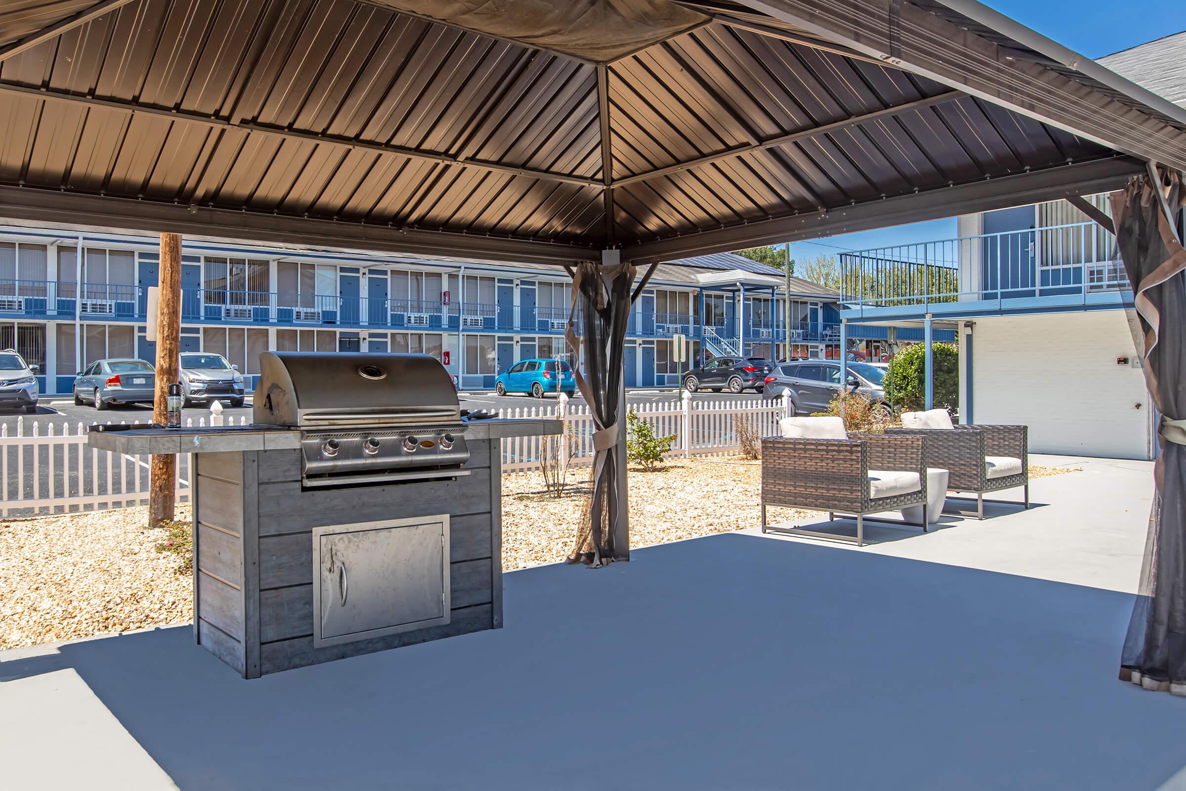 A shaded outdoor grilling area featuring a stainless steel barbecue grill and comfortable seating. The space is designed for relaxation and cooking, with a landscaped ground and a view of nearby parked cars and a building in the background.