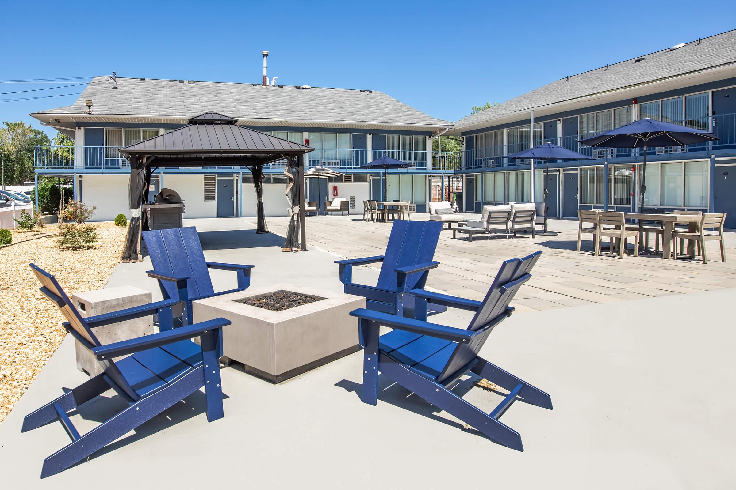 A well-lit outdoor area featuring comfortable blue Adirondack chairs arranged around a fire pit. In the background, a modern two-story building with balconies, along with a gazebo and patio furniture under umbrella shade. The scene is set against a clear blue sky, creating a welcoming atmosphere for relaxation.