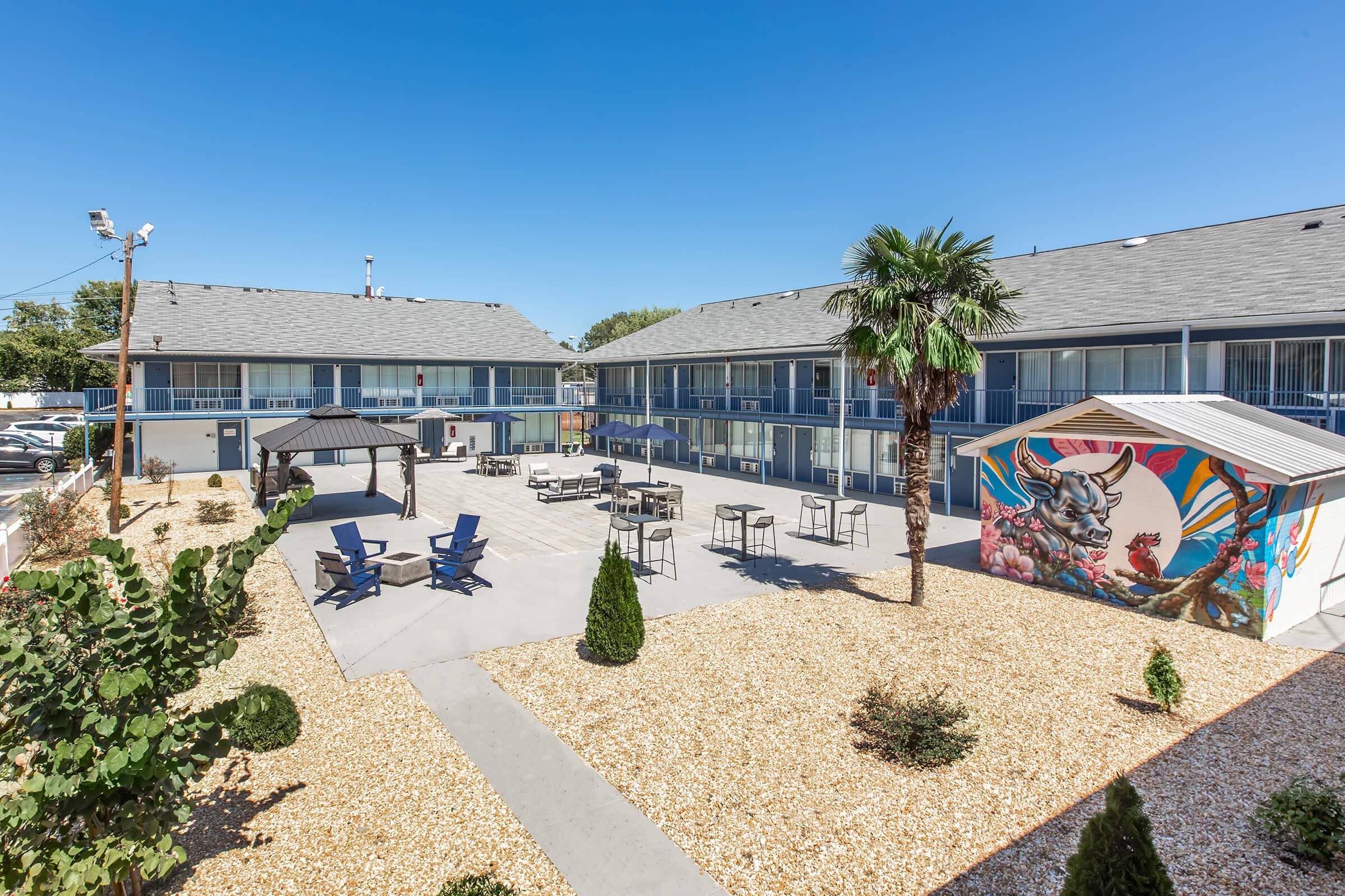 View of a hotel courtyard featuring a patio area with tables and chairs, a gazebo, and a colorful mural on a building. Surrounding the courtyard are palm trees and landscaped gravel areas. The hotel rooms are visible in the background, creating a welcoming outdoor space for guests.