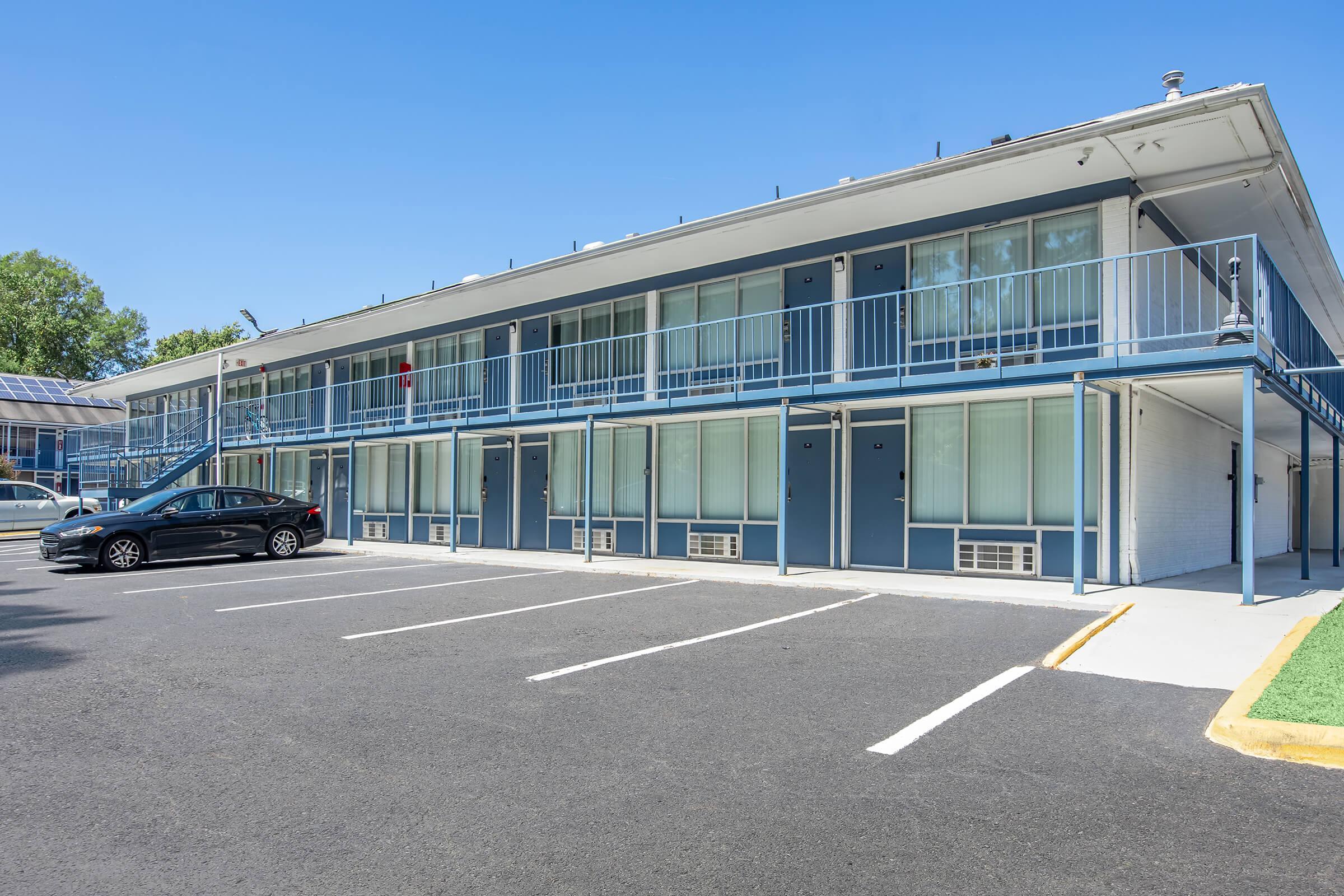 Two-story motel building with a blue and white exterior. Each unit has a balcony and large windows. A black car is parked in front of the building on a paved parking lot. The sky is clear and blue, and there is a small green area with grass alongside the parking lot.
