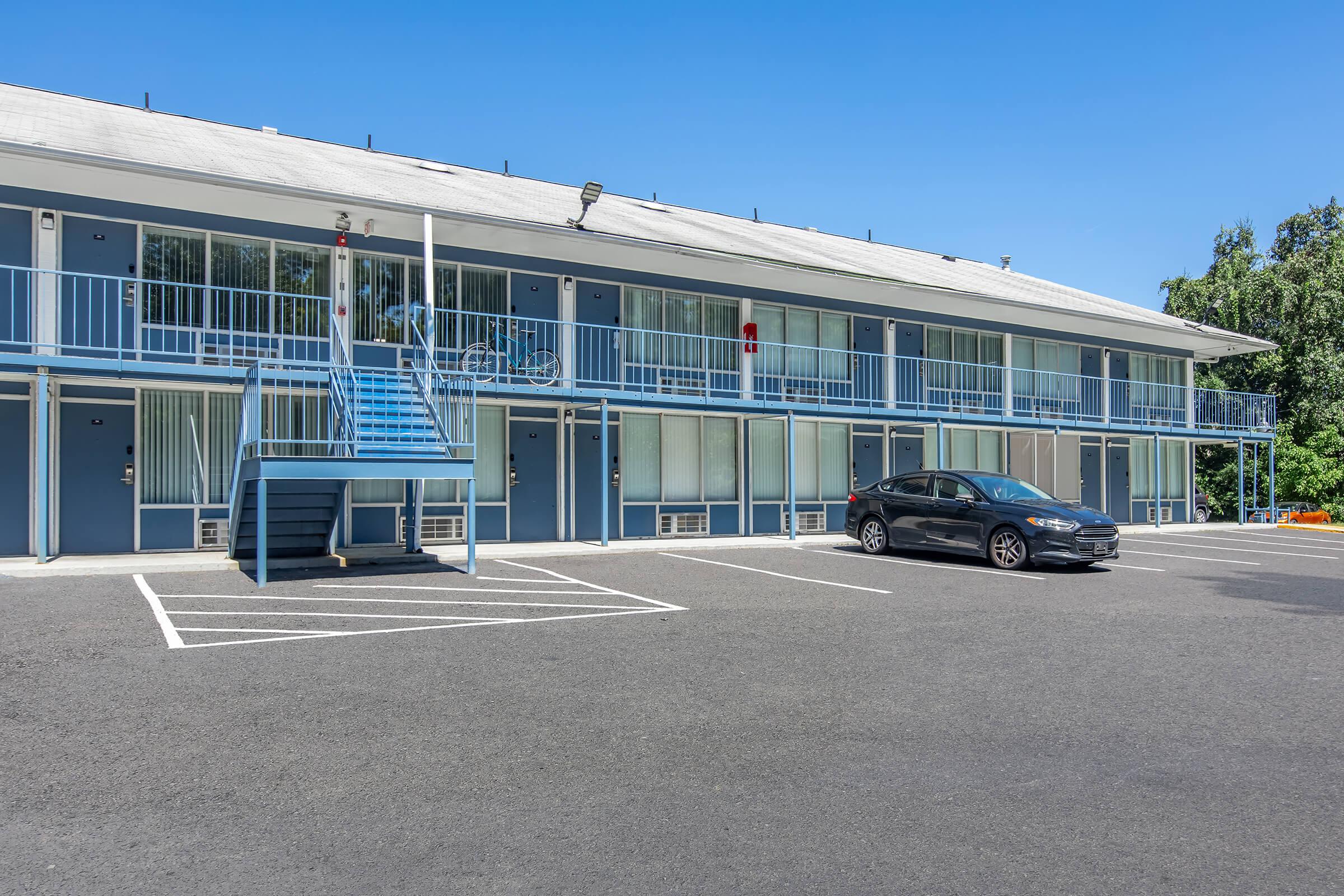 A two-story motel with a bright blue exterior. The building features several doors and a central staircase, with a bicycle leaning against the railing. A black car is parked in the foreground, while the surroundings include clear blue skies and greenery in the background.