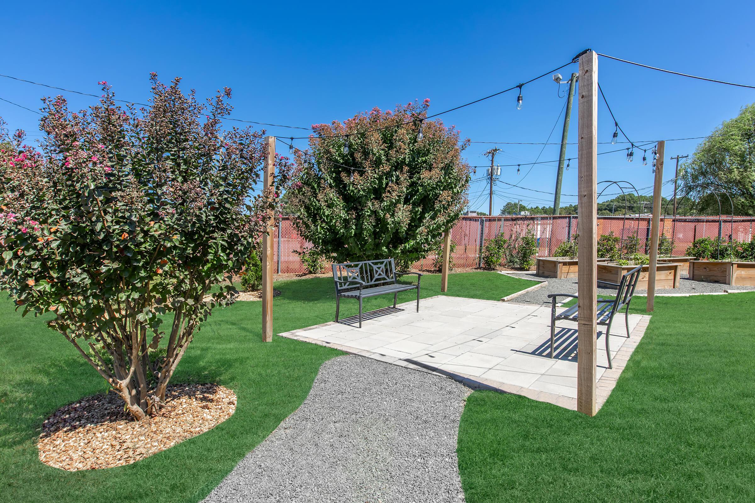 A landscaped outdoor area featuring a stone pathway leading to a seating area with benches. Surrounding the space are flowering plants and trees, with strings of decorative lights overhead. The scene is bathed in daylight, showcasing a bright blue sky.