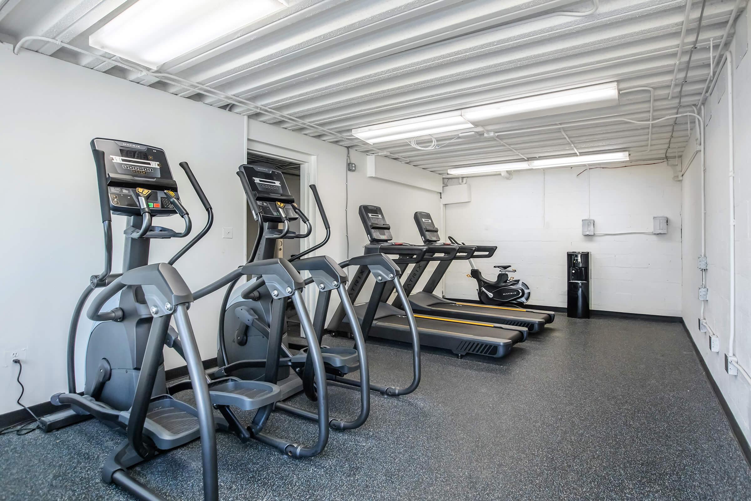 A fitness room with three treadmills and an elliptical machine, featuring a modern design and rubber flooring. The walls are painted white, and there are fluorescent lights above, providing bright illumination. A wall outlet is visible, and the space appears well-maintained and spacious.