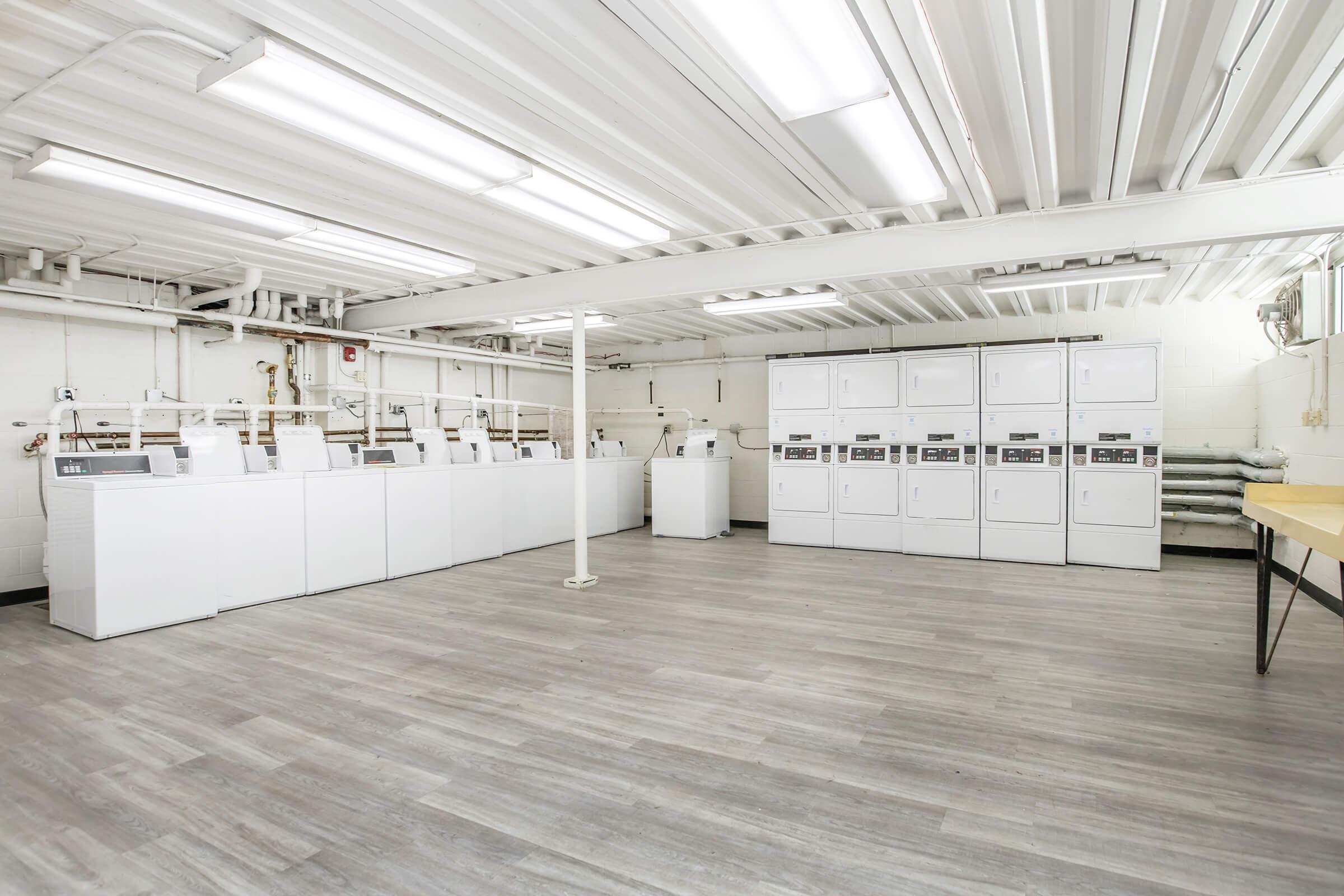 A clean and spacious laundry room featuring multiple white washing machines and dryers aligned against the wall. The floor is covered with light-colored laminate, and the ceiling has white fluorescent lights. The atmosphere is bright and organized, ideal for doing laundry.