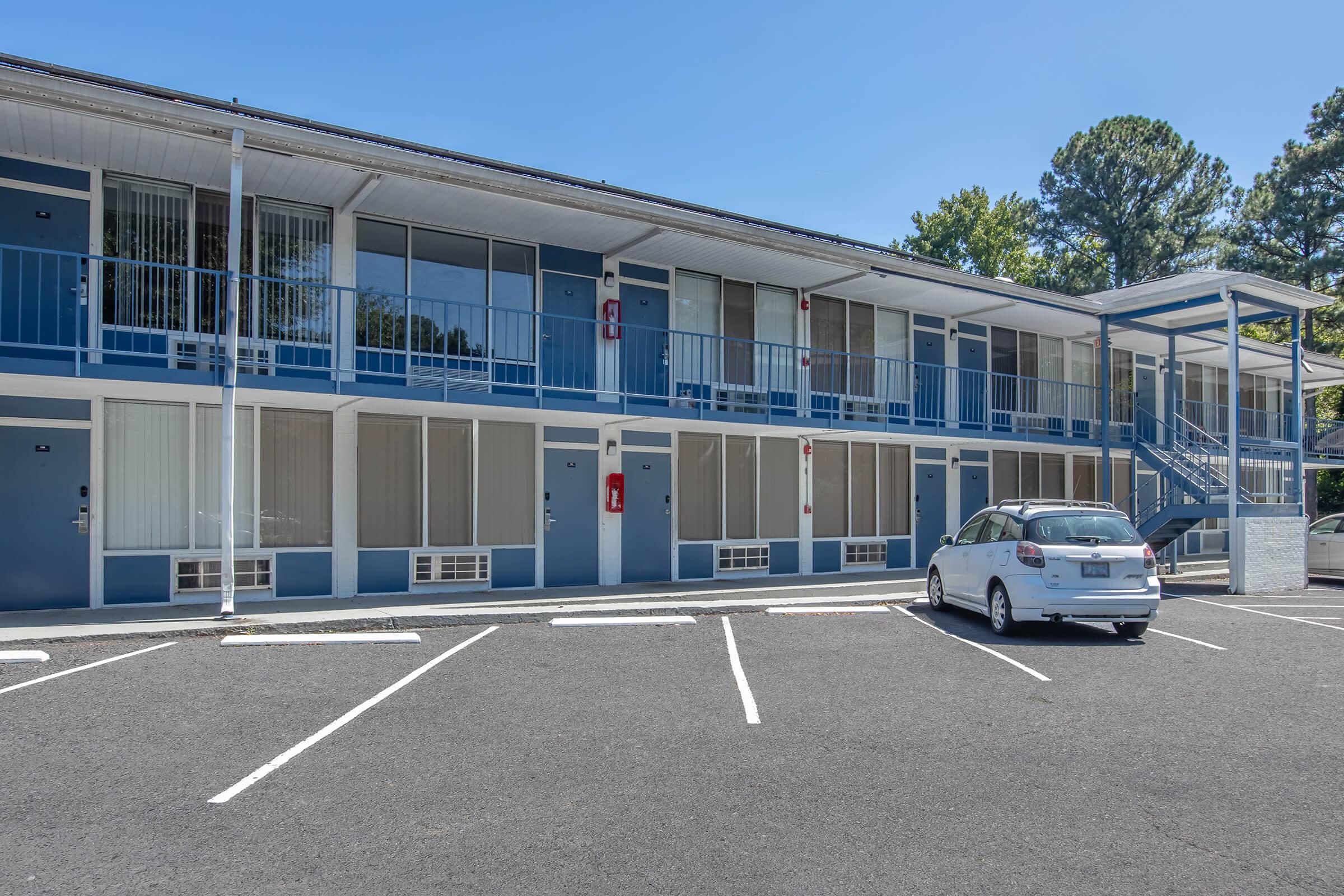 Exterior view of a two-story motel with blue and white walls. The building features several rooms, each with large windows and balconies. A white car is parked in front, and there is a staircase leading to the upper floor. The surrounding area is clear, with trees in the background and parking spaces visible.