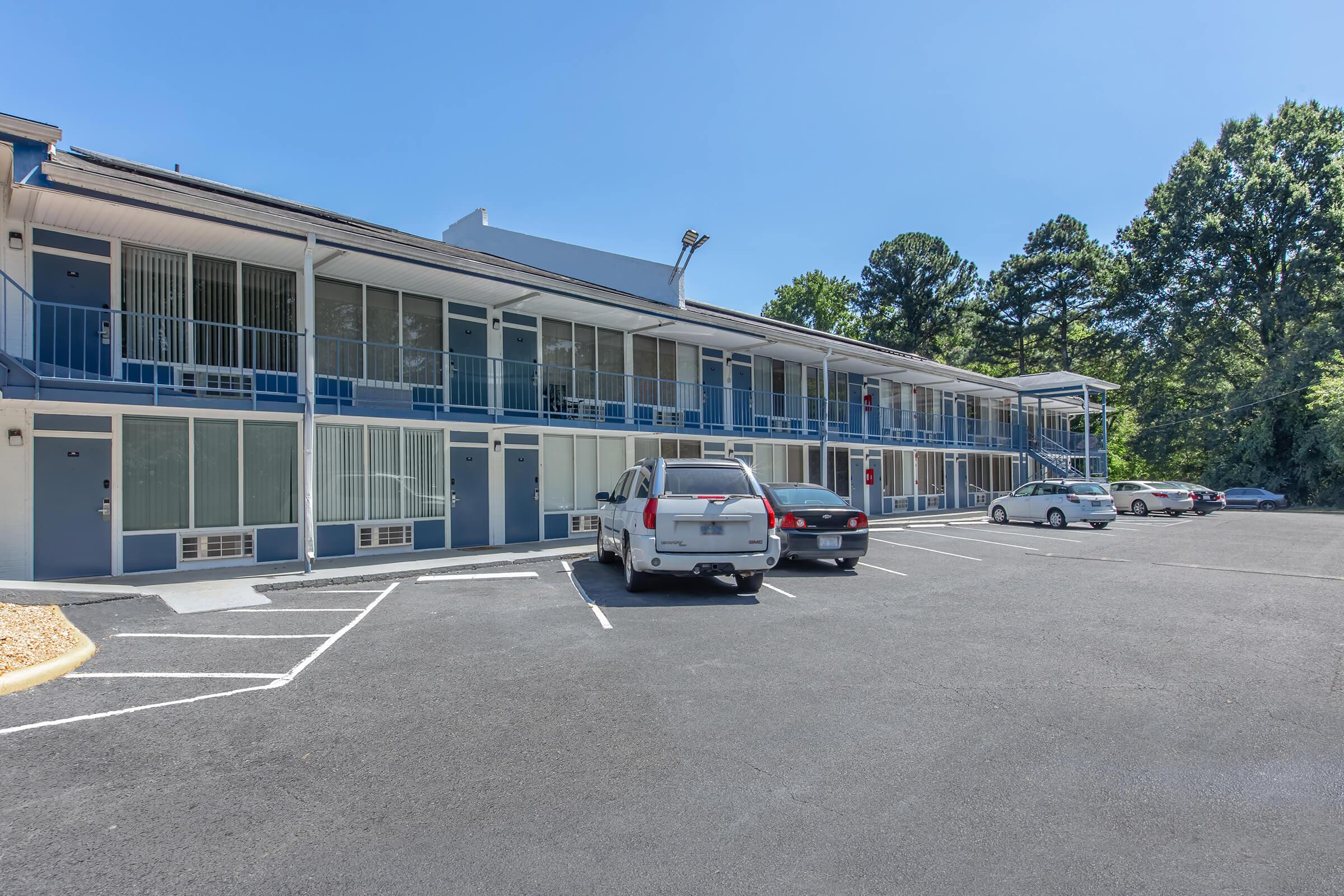 A low-rise motel with blue and white exterior, featuring multiple rooms with balconies. Several cars are parked in the lot in front of the building, surrounded by trees. Clear blue sky overhead indicates a sunny day.