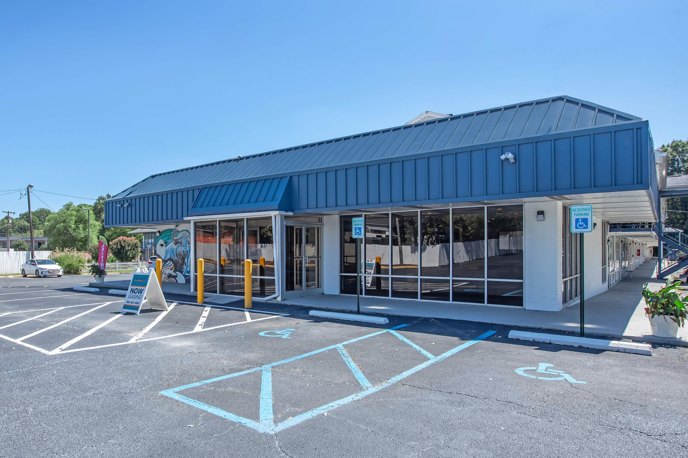A modern single-story commercial building with a blue metal roof and large windows. The exterior features a mural on one side, and there are several parking spaces, including accessible spots. A sign outside indicates the business and there are landscaped areas with plants. Clear blue sky in the background.