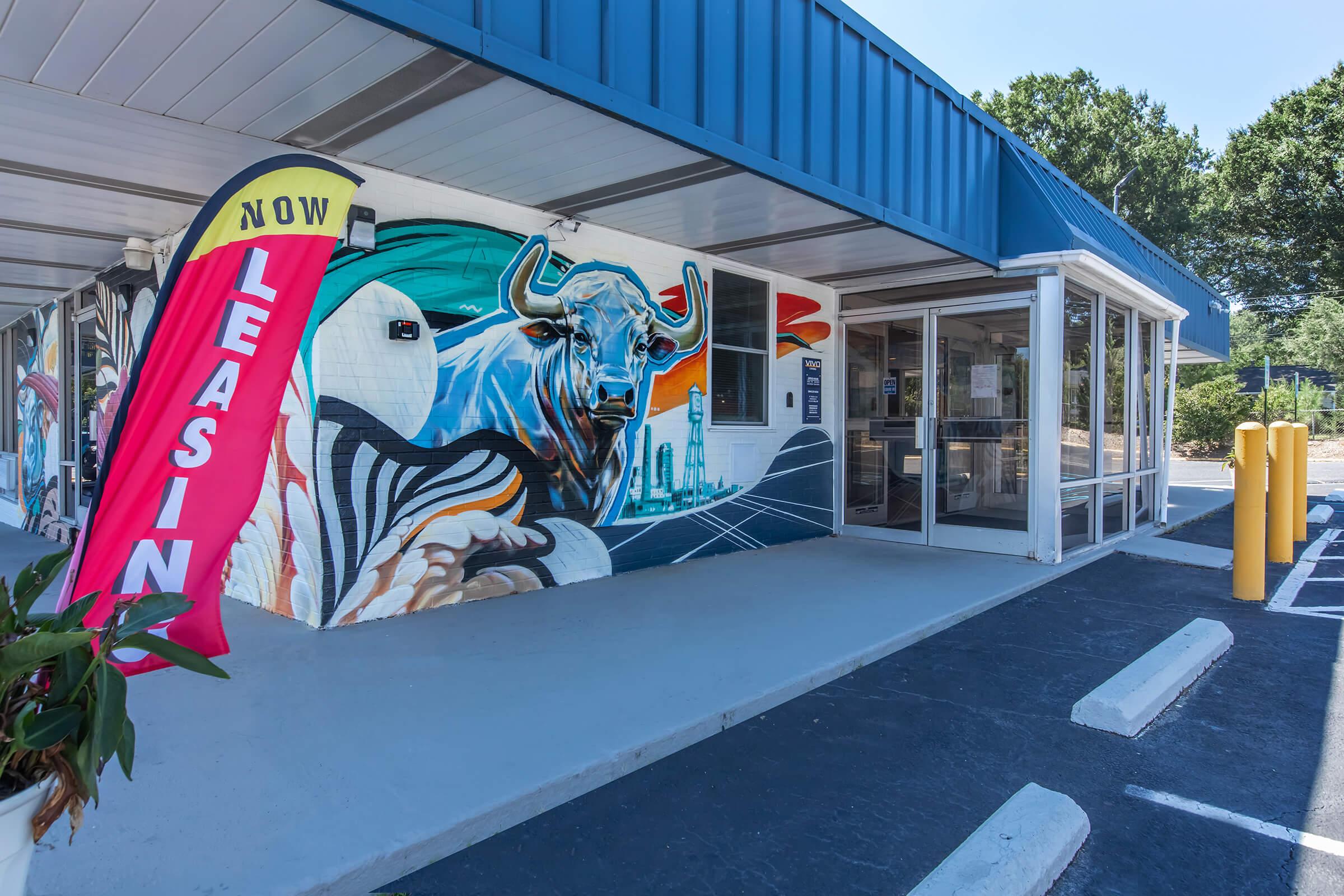 A vibrant mural featuring a bull and abstract designs adorns the exterior wall of a commercial building. In front, a red "Now Leasing" banner flutters, indicating available rental space. The entrance is visible, with a contemporary design and surrounding parking spaces. Green foliage adds a touch of nature to the scene.