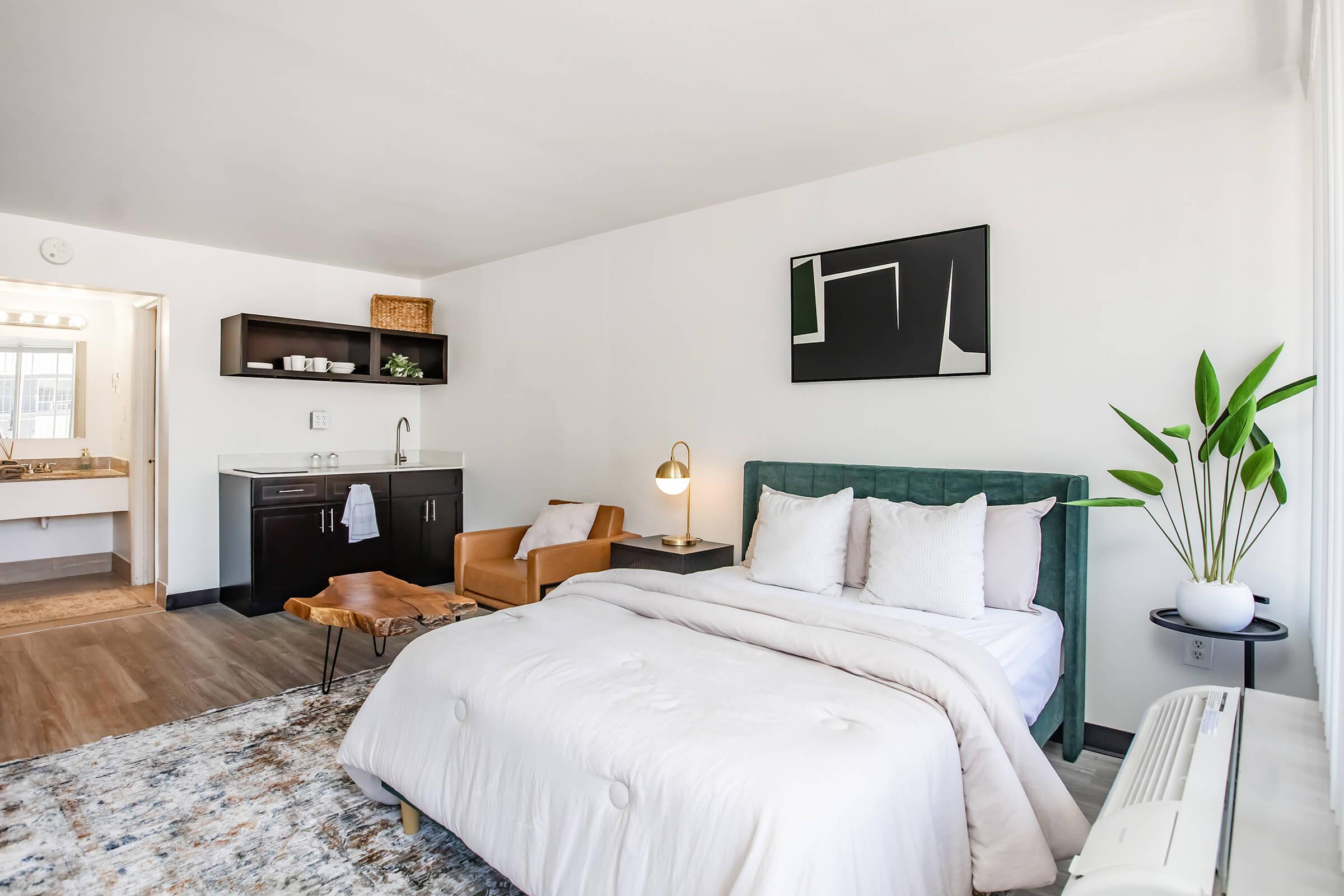 A cozy bedroom featuring a green upholstered bed with white bedding, a stylish armchair, a modern nightstand with a lamp, and a wall art piece. There's a kitchenette with dark cabinetry in the background and a bathroom visible through an open door, along with indoor plants for a fresh touch.
