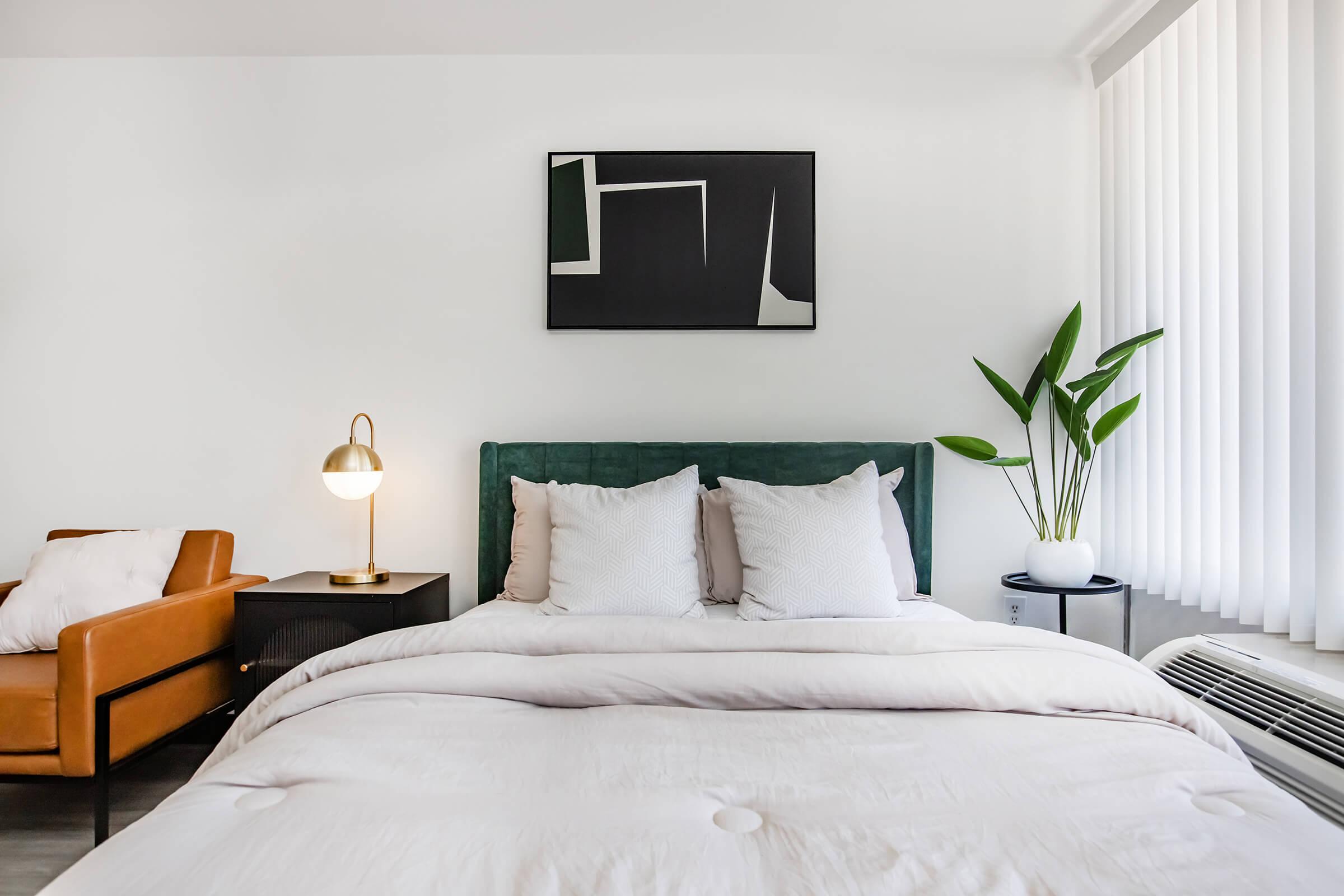 A modern bedroom featuring a green upholstered headboard with white pillows on a bed, a stylish lamp on a bedside table, a comfortable brown armchair, and a tall plant near the window with vertical blinds. An abstract painting hangs above the bed, enhancing the contemporary decor.