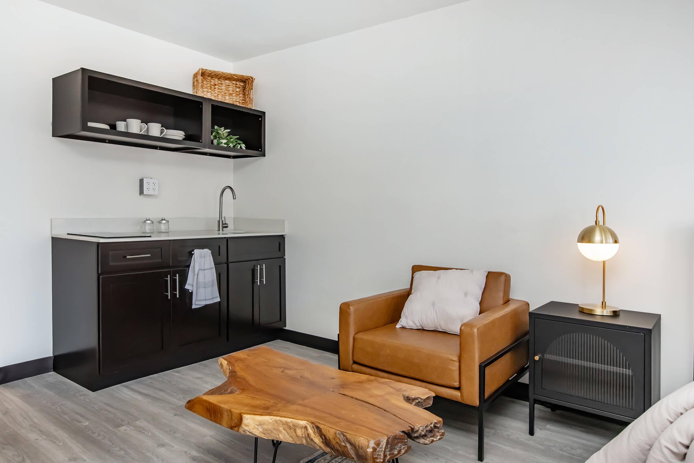 A modern, minimalist living space featuring a brown leather armchair, a wooden coffee table, and a sleek kitchenette with dark cabinetry. There’s a light-colored wall, a small sink, and decorative items on the shelf. A stylish lamp is placed beside the chair, enhancing the cozy ambiance.