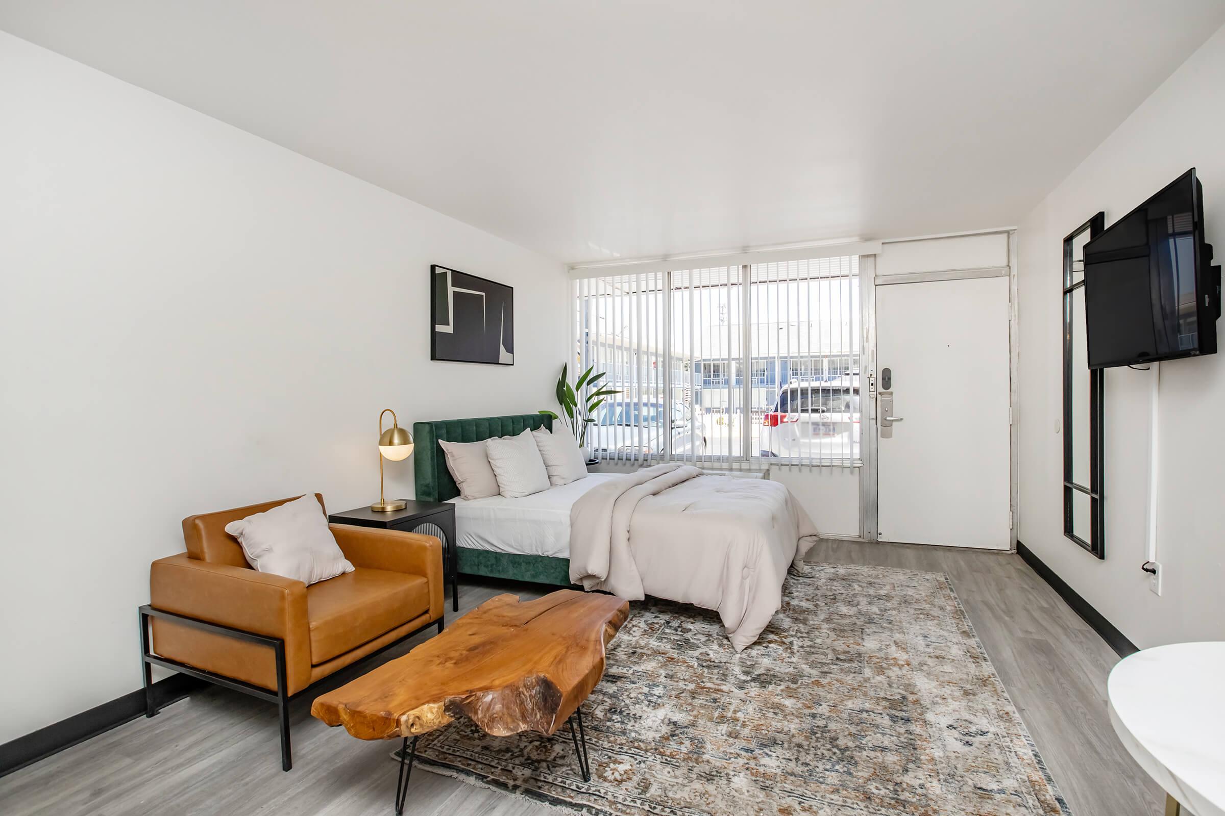A modern, minimally designed hotel room featuring a green bed with gray bedding, a wooden coffee table, an orange armchair, and a large window with blinds. A wall-mounted TV is visible, along with a small plant and abstract wall art, creating a cozy and inviting atmosphere.