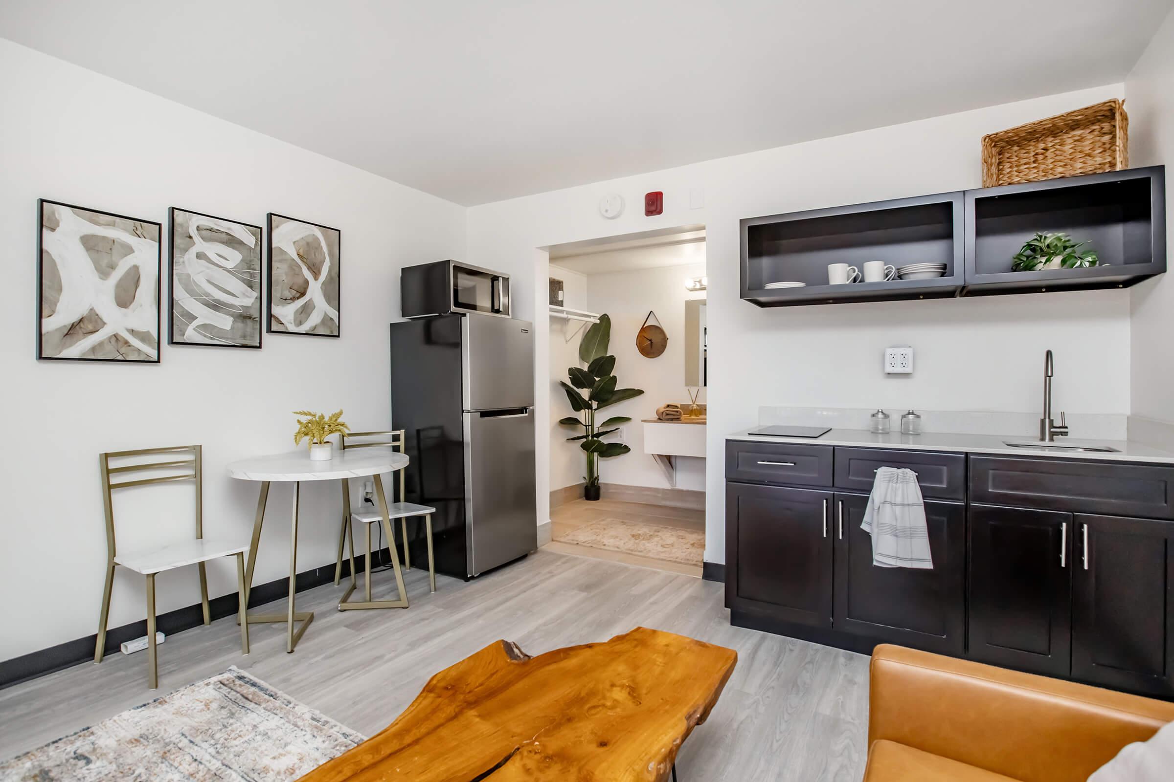 A modern kitchen and dining area featuring a small round table with two chairs, a sleek black refrigerator, and dark cabinetry. Decor includes three framed artworks on the wall, a plant in the corner, and a wooden coffee table in the foreground, enhancing the contemporary design feel.