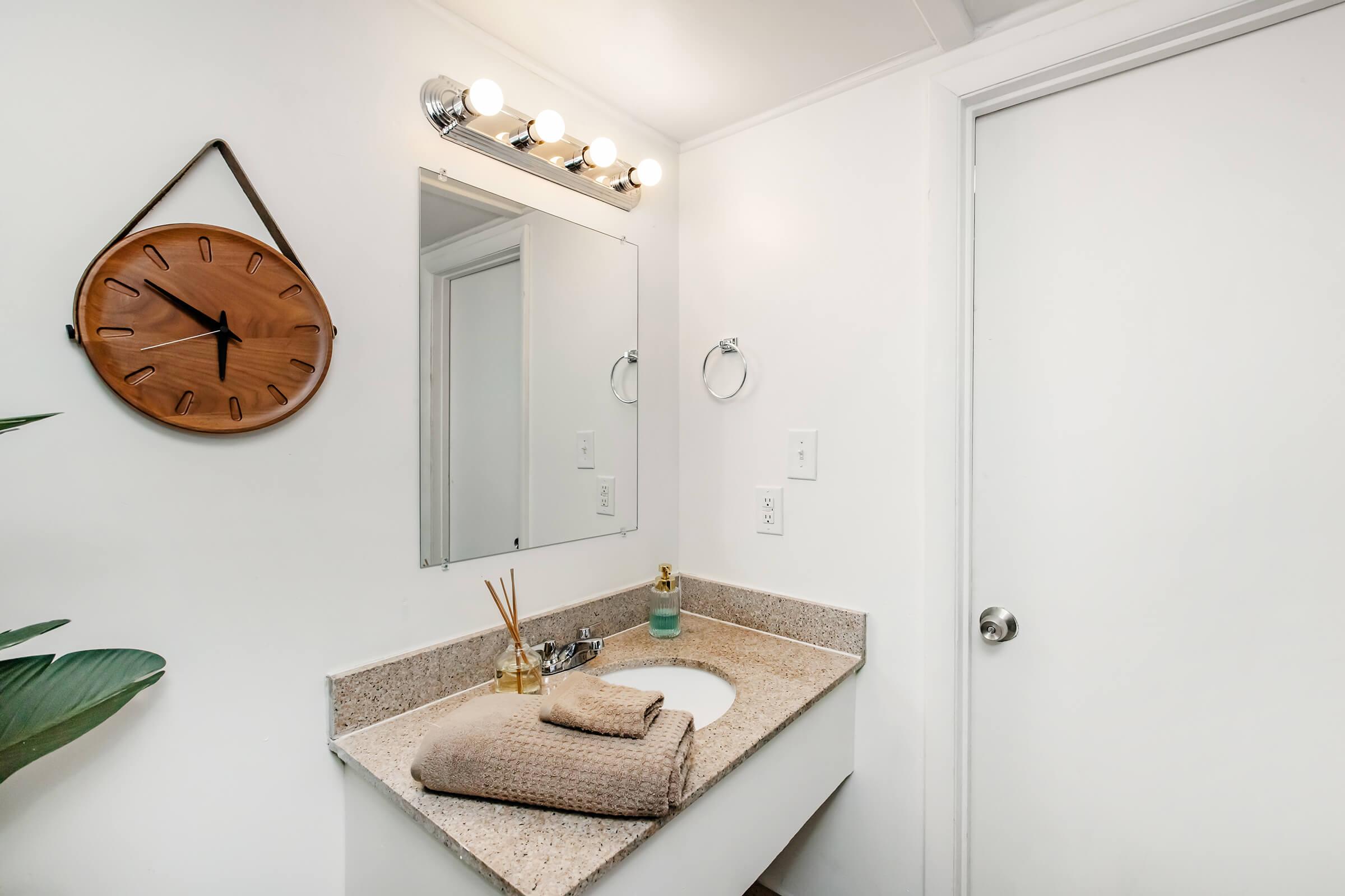 A modern bathroom featuring a light-colored countertop with a sink, a decorative wooden clock on the wall, a mirror, and a towel neatly folded beside a small plant. Soft lighting fixtures above the mirror enhance the bright, clean aesthetic of the space.