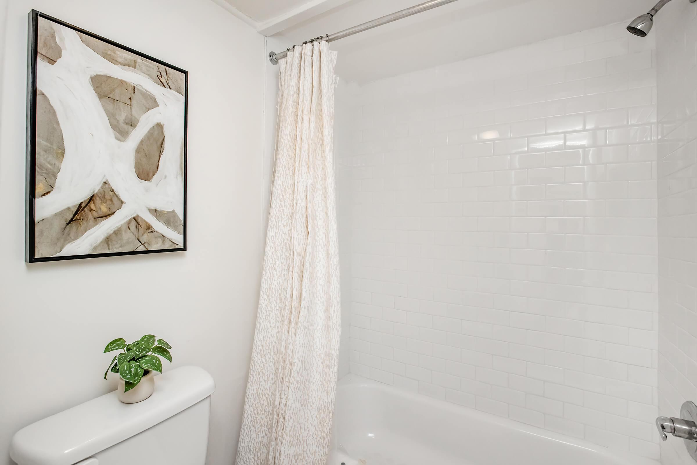 A clean and modern bathroom featuring a white tiled shower area and a bathtub. There is a light-colored curtain partially drawn, and a small green plant sits on top of the toilet. A framed abstract artwork with light and dark colors adorns the wall, adding a decorative touch to the space.