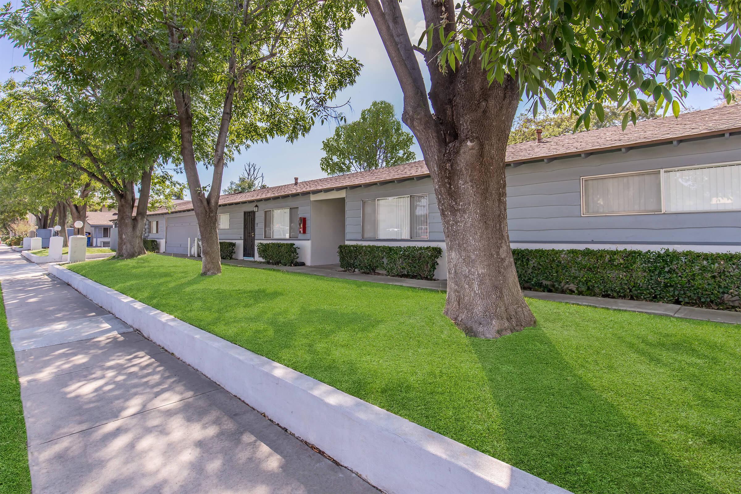 A quiet residential street lined with well-maintained, single-story gray apartment buildings. Lush green lawns and trees create a serene atmosphere. Sidewalks are clear, and the surroundings suggest a pleasant, suburban setting with bright sunlight filtering through the leaves.