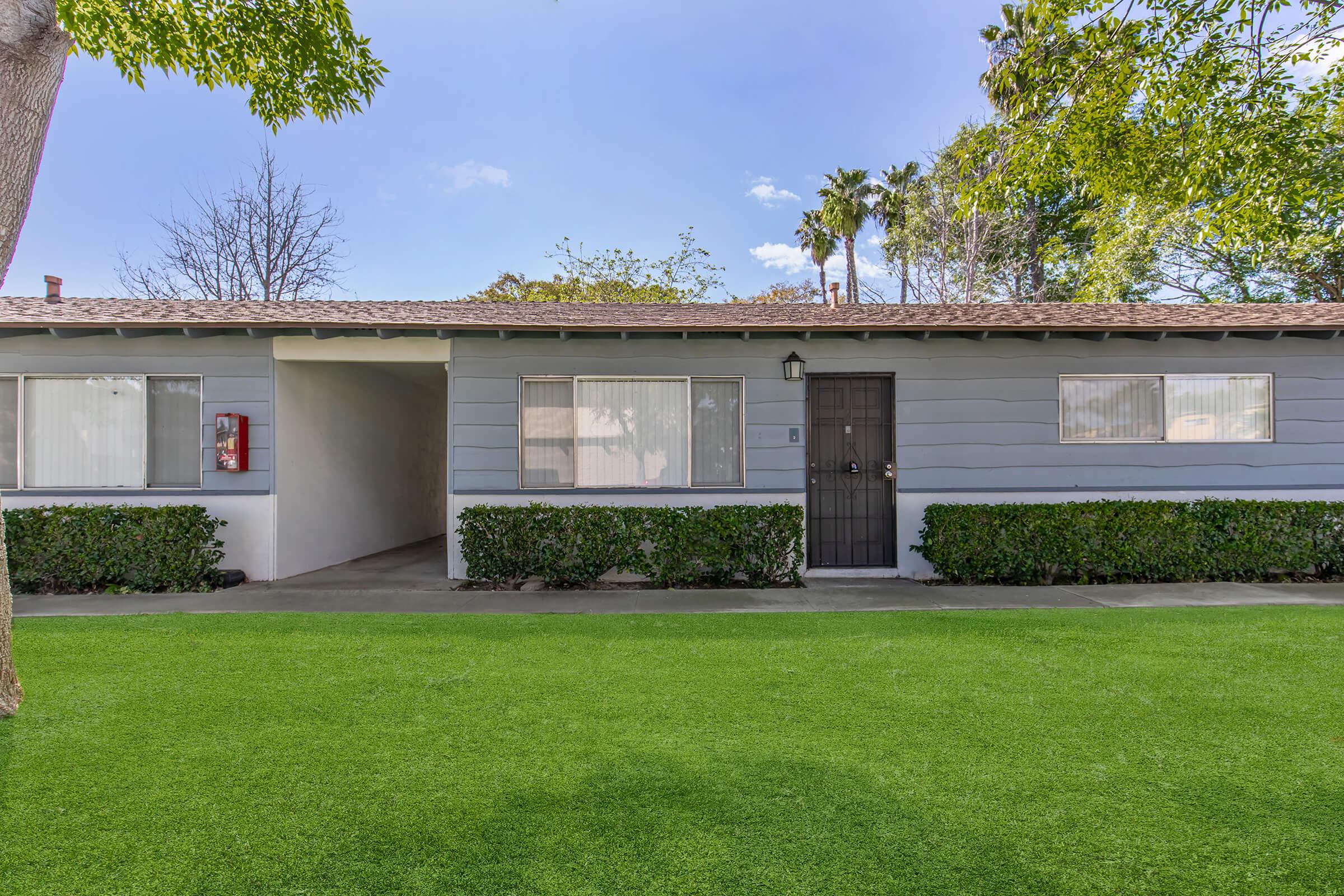 A single-story residential building with a gray exterior, featuring multiple windows and a black door. The front yard is covered in green grass, with neat hedges lining the walkway. Trees and palm trees are visible in the background against a clear blue sky.