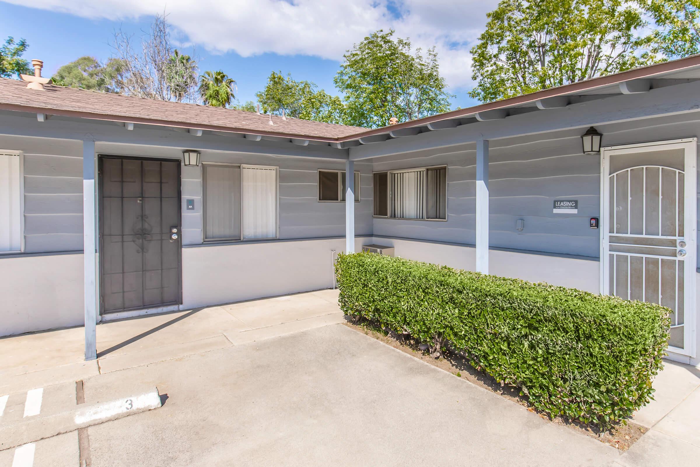 A view of a single-story building with gray wooden siding. The entrance features a dark door and a small front porch area. Nearby, a neatly trimmed hedge lines the front, and a paved parking area is visible. Trees and blue sky with clouds can be seen in the background.