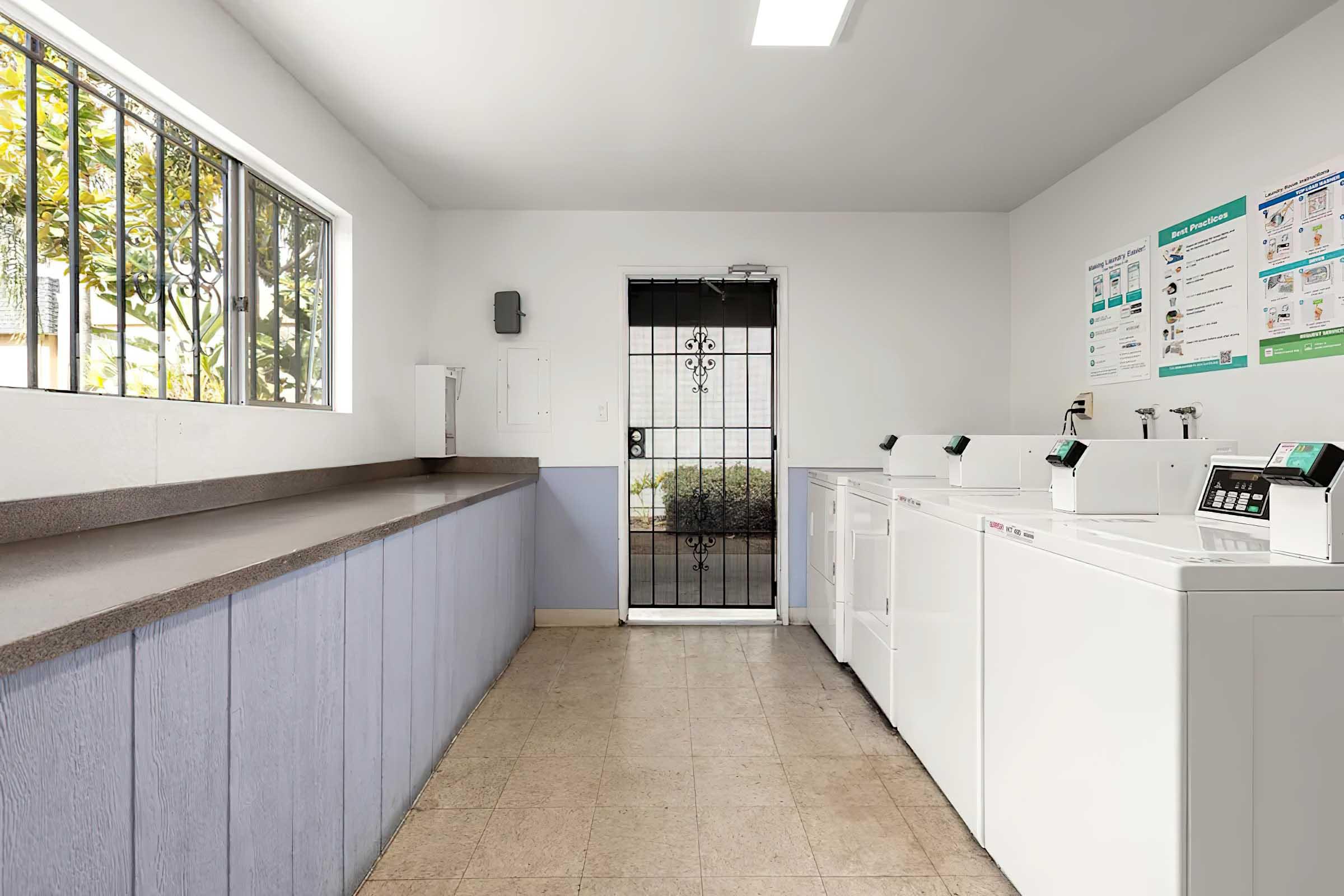 Laundry room with several white washing machines lined up against a wall. There is a long countertop on one side and a door with a security gate leading outside. Natural light comes through a window, and instructional posters are mounted on the wall. The space appears clean and well-maintained.