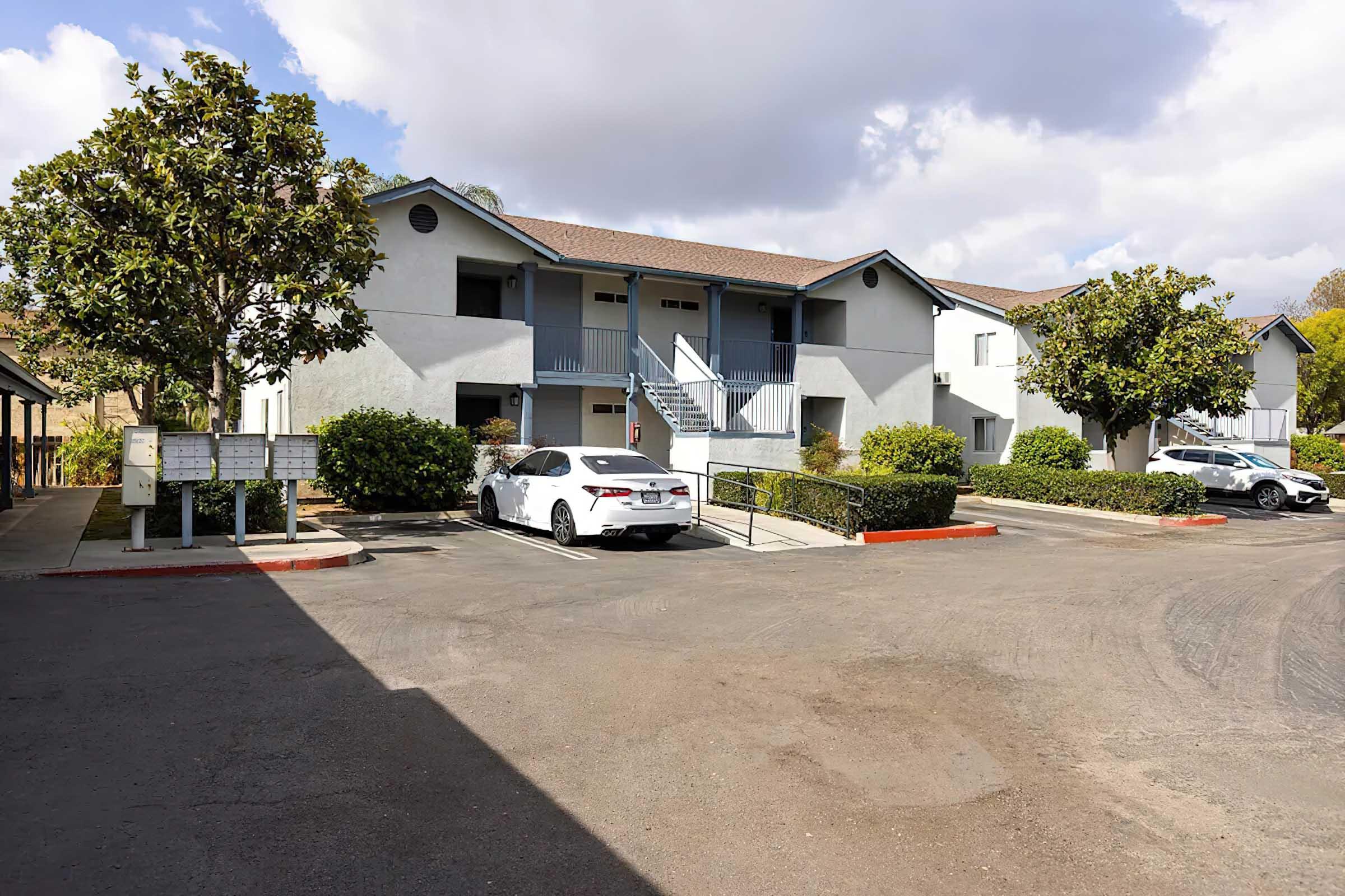 A residential complex featuring beige two-story buildings with blue accents. The foreground shows a parking area with parked cars and neatly trimmed hedges. A few mailboxes are visible, and the scene is set under a partly cloudy sky.