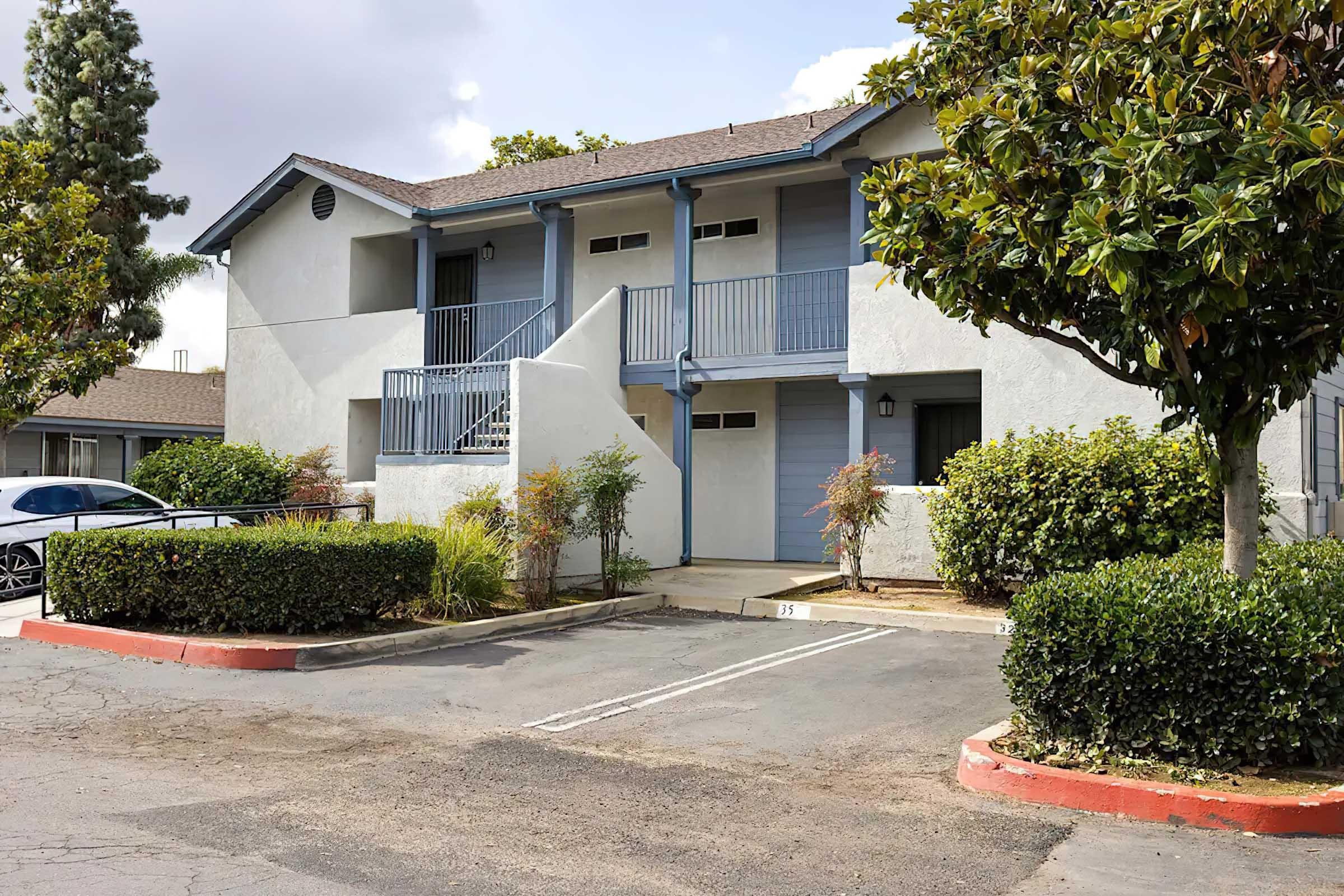 A multi-unit residential apartment building with a light-colored exterior. The structure features a staircase leading to upper-level units and is surrounded by greenery, including bushes and small trees. Two parking spaces are visible in front of the building, which is set against a partly cloudy sky.