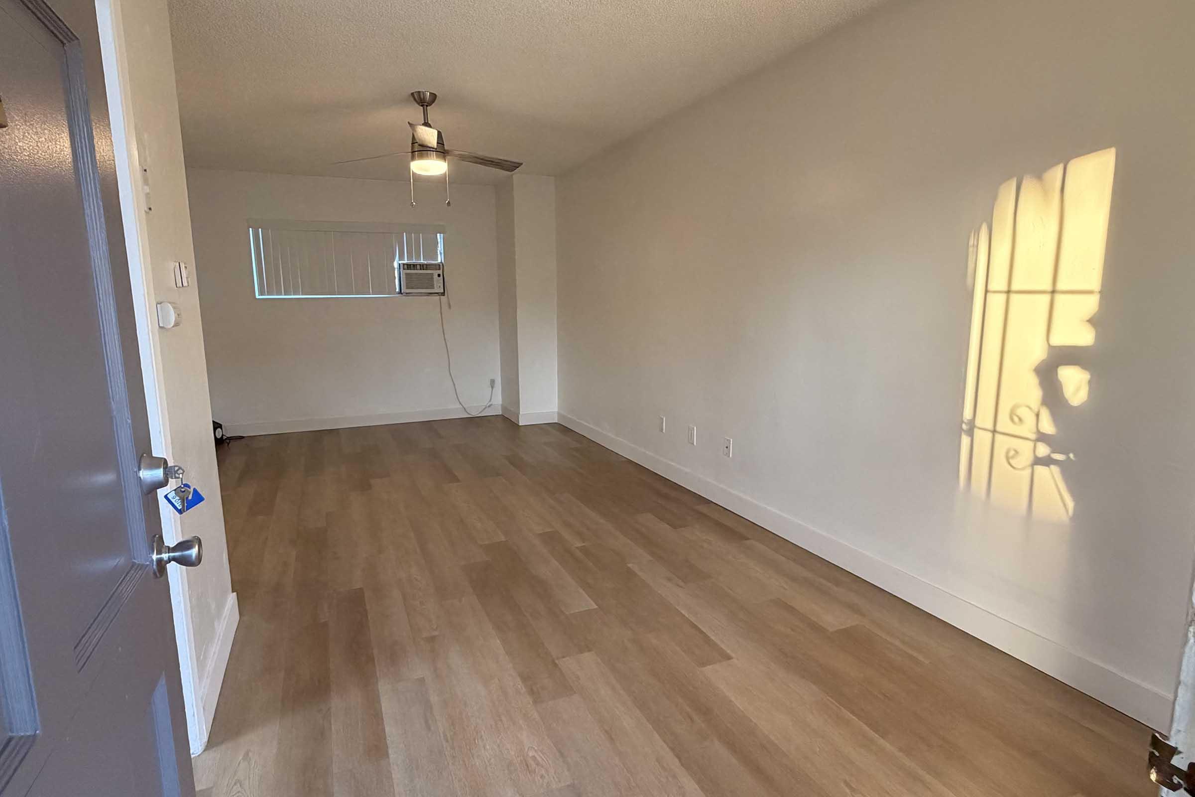 Interior view of a vacant room with light wood flooring, a ceiling fan, and a window casting a shadow on the wall. The space is well-lit, featuring a neutral color palette and minimal furnishings, creating a spacious and clean environment.