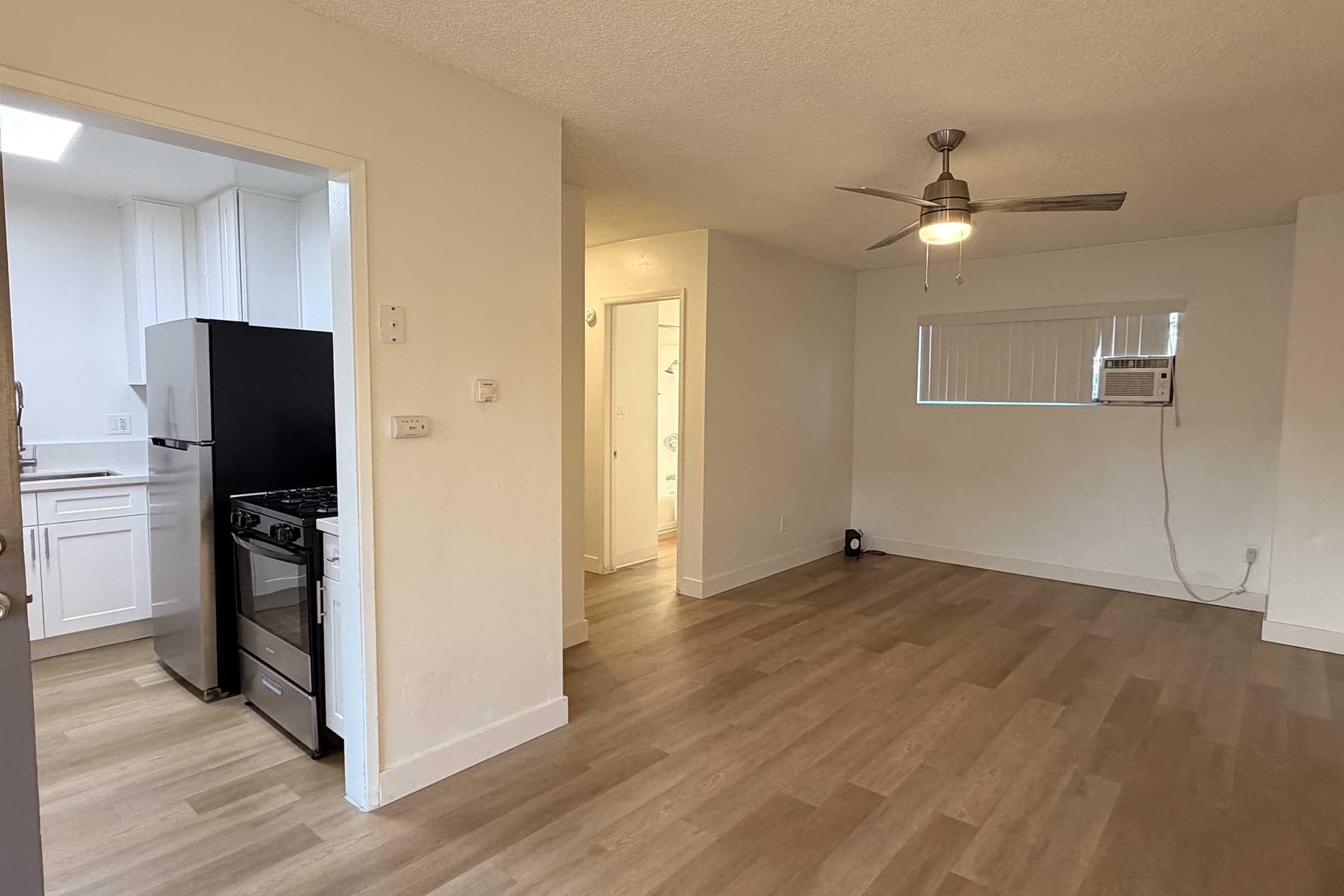 A well-lit, open-concept living area featuring a kitchen with a black refrigerator and a gas stove on the left. The space has light-colored walls and wood flooring, with a ceiling fan and a window facing the living area. An adjacent door leads to another room, providing a cozy and inviting atmosphere.