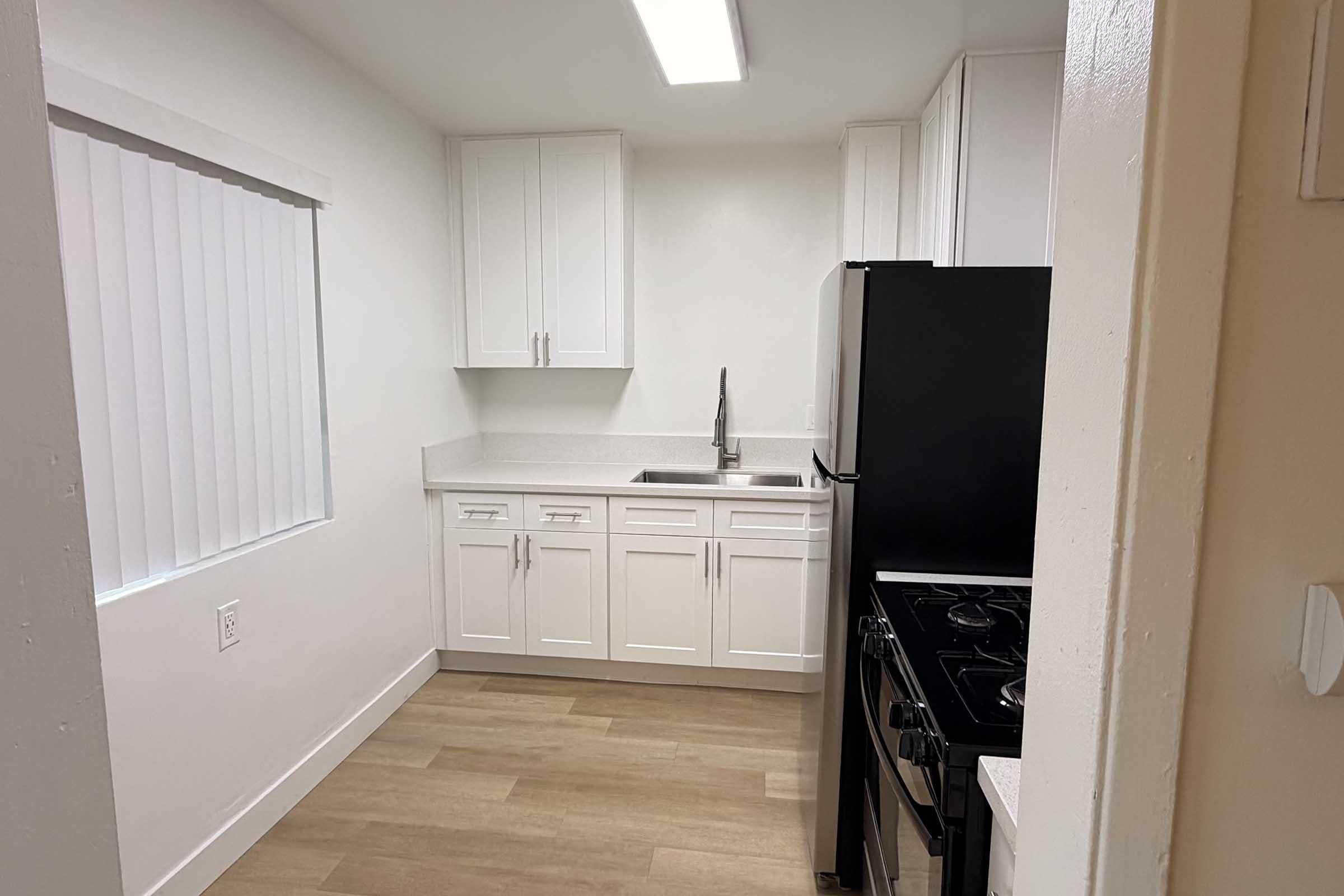 A modern kitchen featuring white cabinets, a black refrigerator, and a stove. The space has a clean, bright appearance with a window partially covered by blinds. The flooring is light wood, and the area is well-lit by overhead lighting.