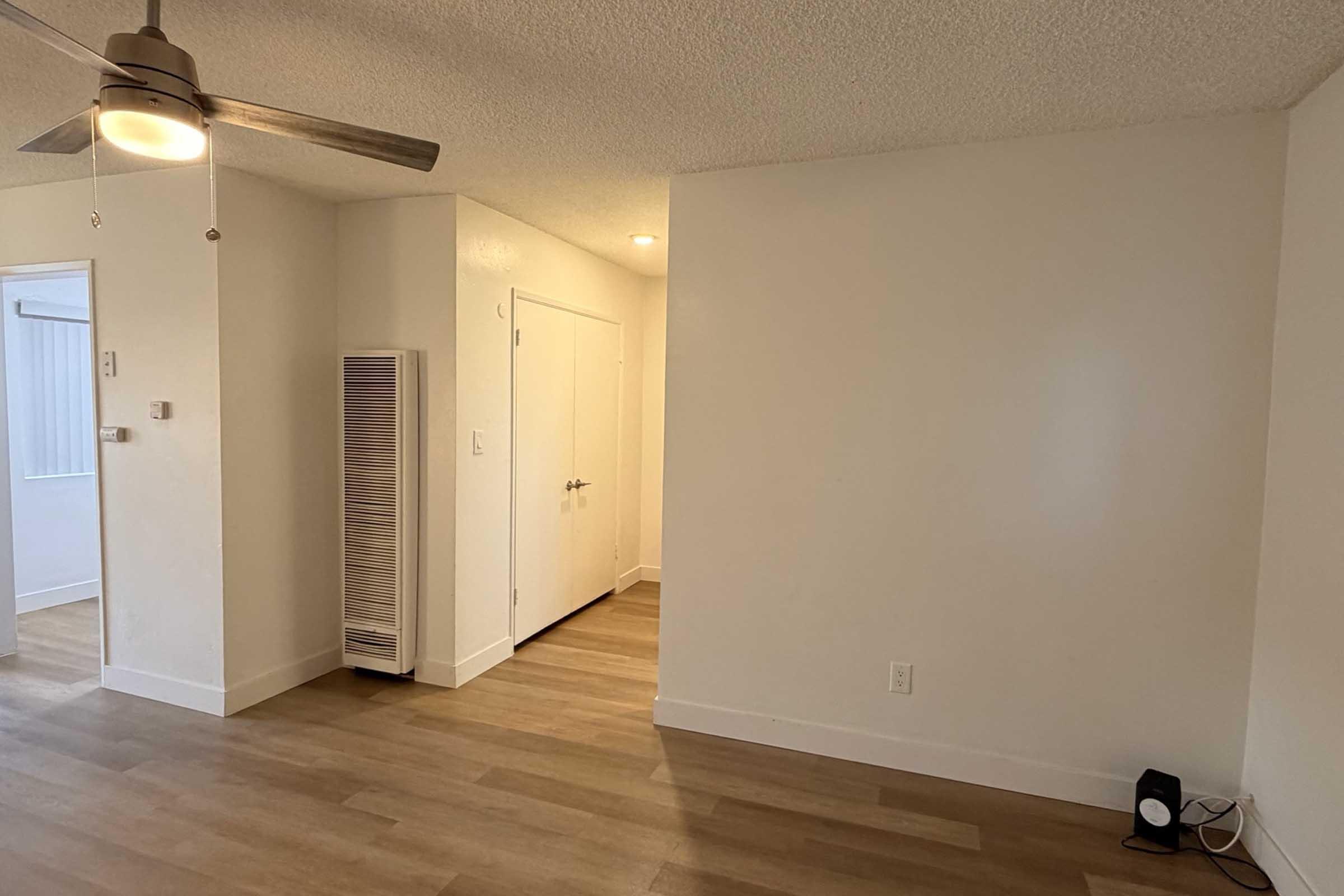 A well-lit, empty living space with light-colored walls and laminate flooring. There is a ceiling fan, a narrow white heater on the wall, and an open doorway leading to another room. In the lower right corner, a small speaker is visible near the wall.