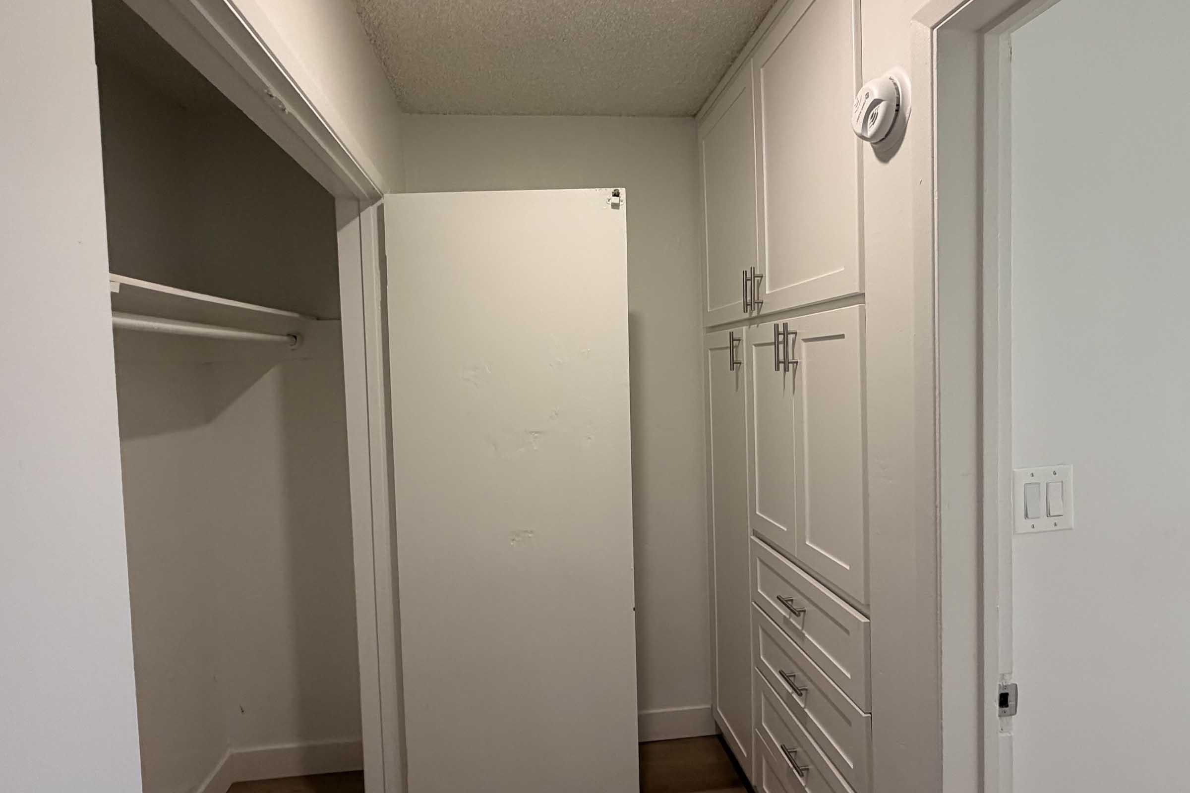 A small, bright room with a white refrigerator on the left, a closet with a horizontal rod for hanging clothes, and light-colored cabinetry on the right featuring multiple drawers and doors. The walls are painted white, and the floor is a light wood finish.