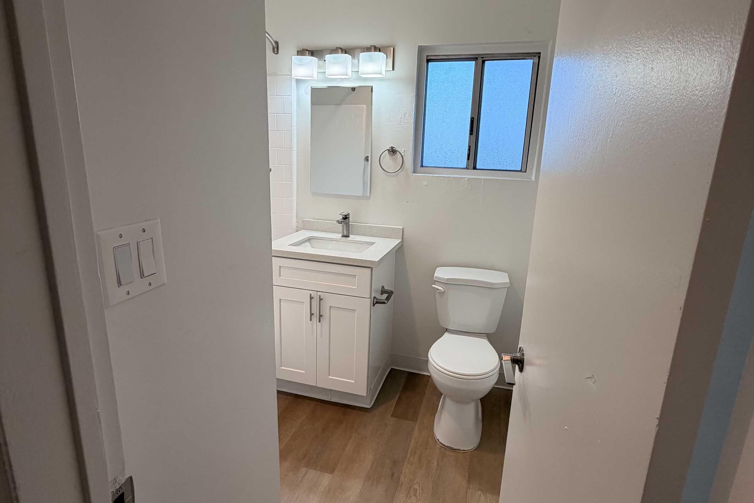 A modern bathroom featuring a white vanity with a sink, a rectangular mirror above, a toilet, and a light fixture with multiple bulbs. There is a window allowing natural light and wooden flooring. The doorway is visible on the left, leading into the bathroom space.