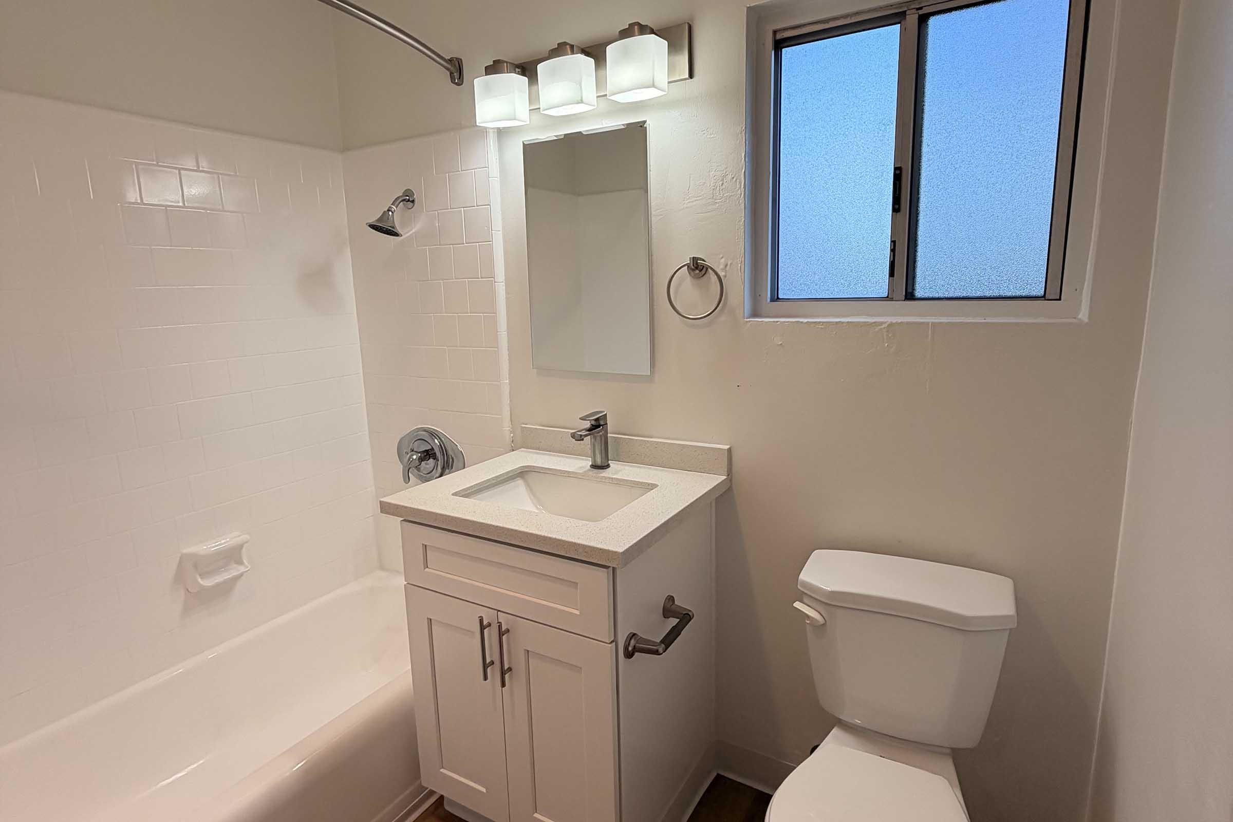 A clean and modern bathroom featuring a white bathtub, a rectangular sink with a sleek countertop, a large mirror above the sink, a showerhead, a towel ring, and a toilet. Natural light comes through a small window with frosted glass, illuminating the space. The walls are painted in a light, neutral tone.