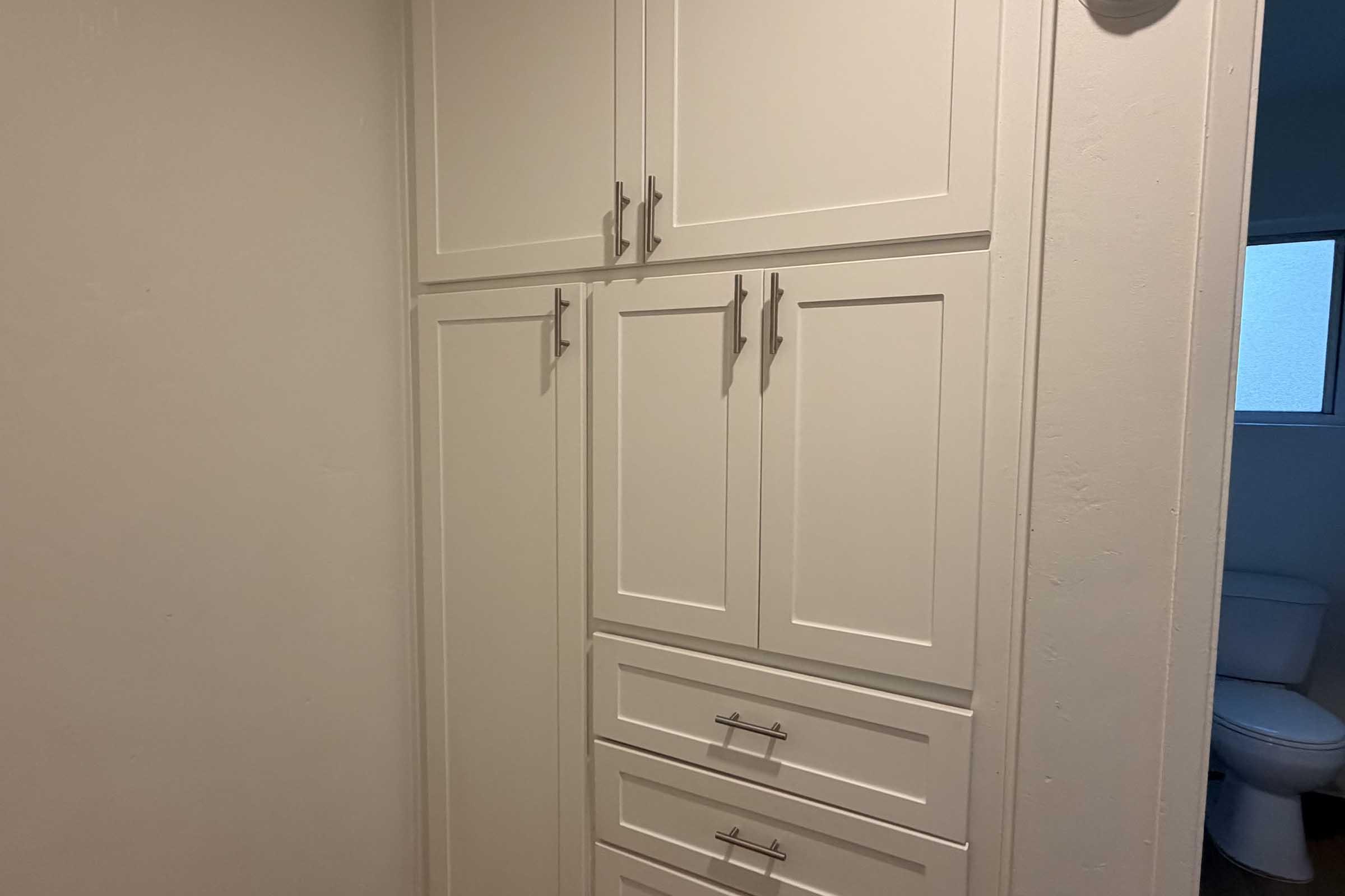A wall of white kitchen cabinets with four upper doors and three lower drawers, featuring modern silver handles. A bathroom can be seen in the background, indicated by a toilet and a window. The walls are painted a light color, creating a clean and bright appearance.