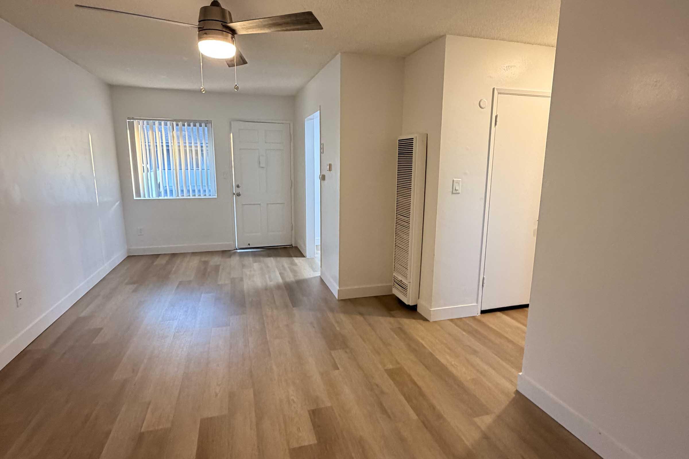 A well-lit interior space featuring light-colored walls and wooden flooring. There is a ceiling fan, a window with vertical blinds, a front door, and a hallway leading to another room. An air conditioning unit is mounted on the wall near the entrance. The area appears clean and recently updated.