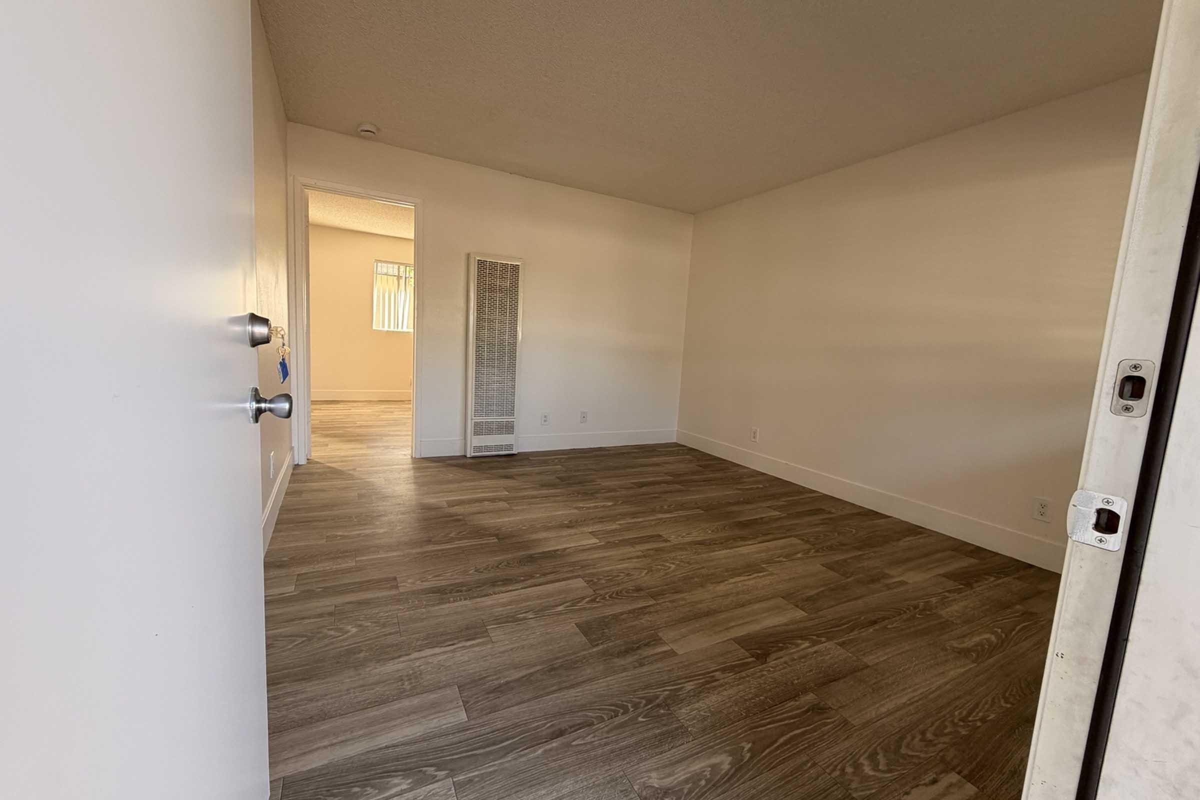 A spacious, empty room with light-colored walls and wooden flooring. A doorway leads to another room, and a vertical heater is visible on the wall. Natural light comes from a window with blinds in the background, creating a bright and open atmosphere.